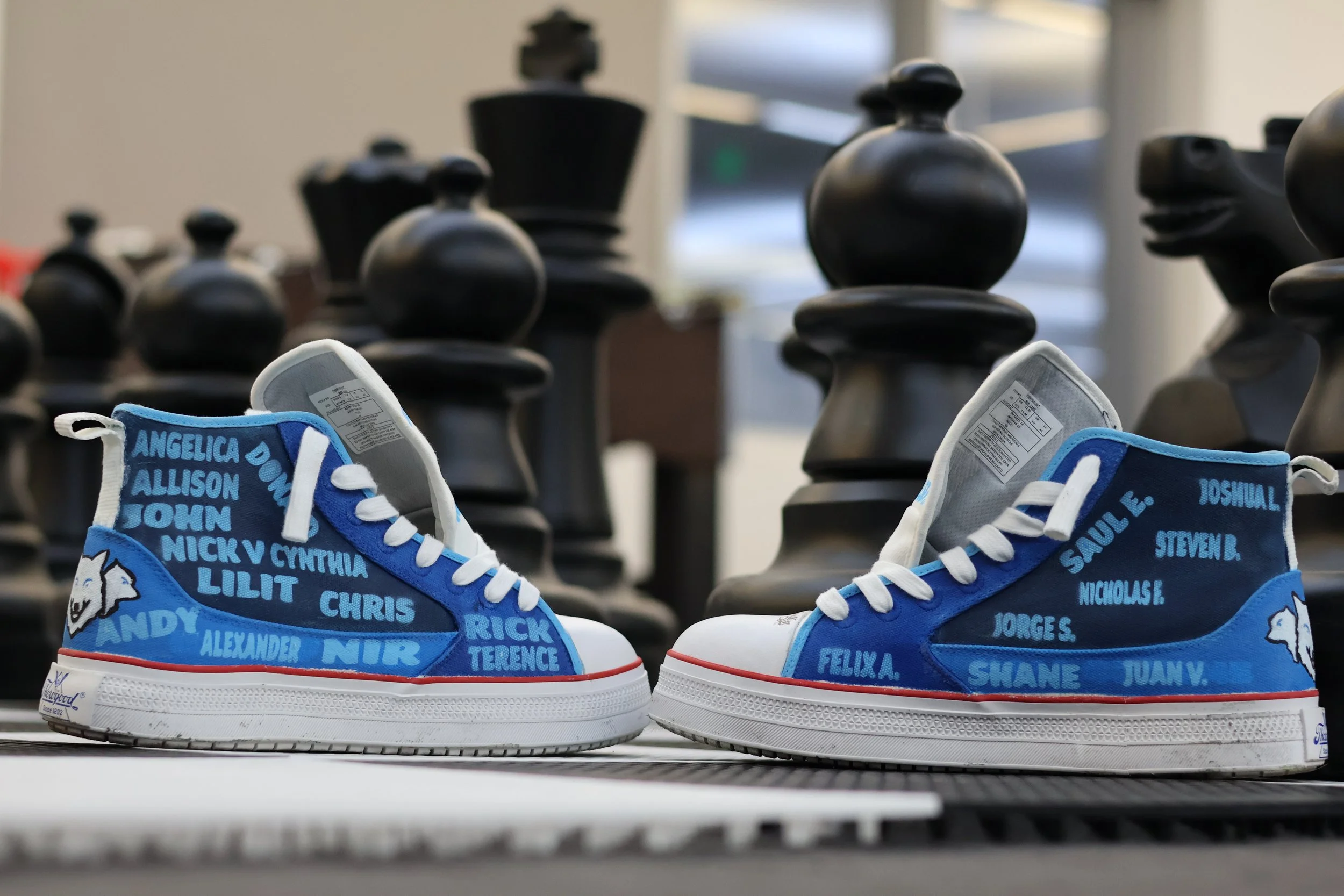 A pair of blue and white sneakers with city names and people's names written on them, placed on a table with large chess pieces in the background.