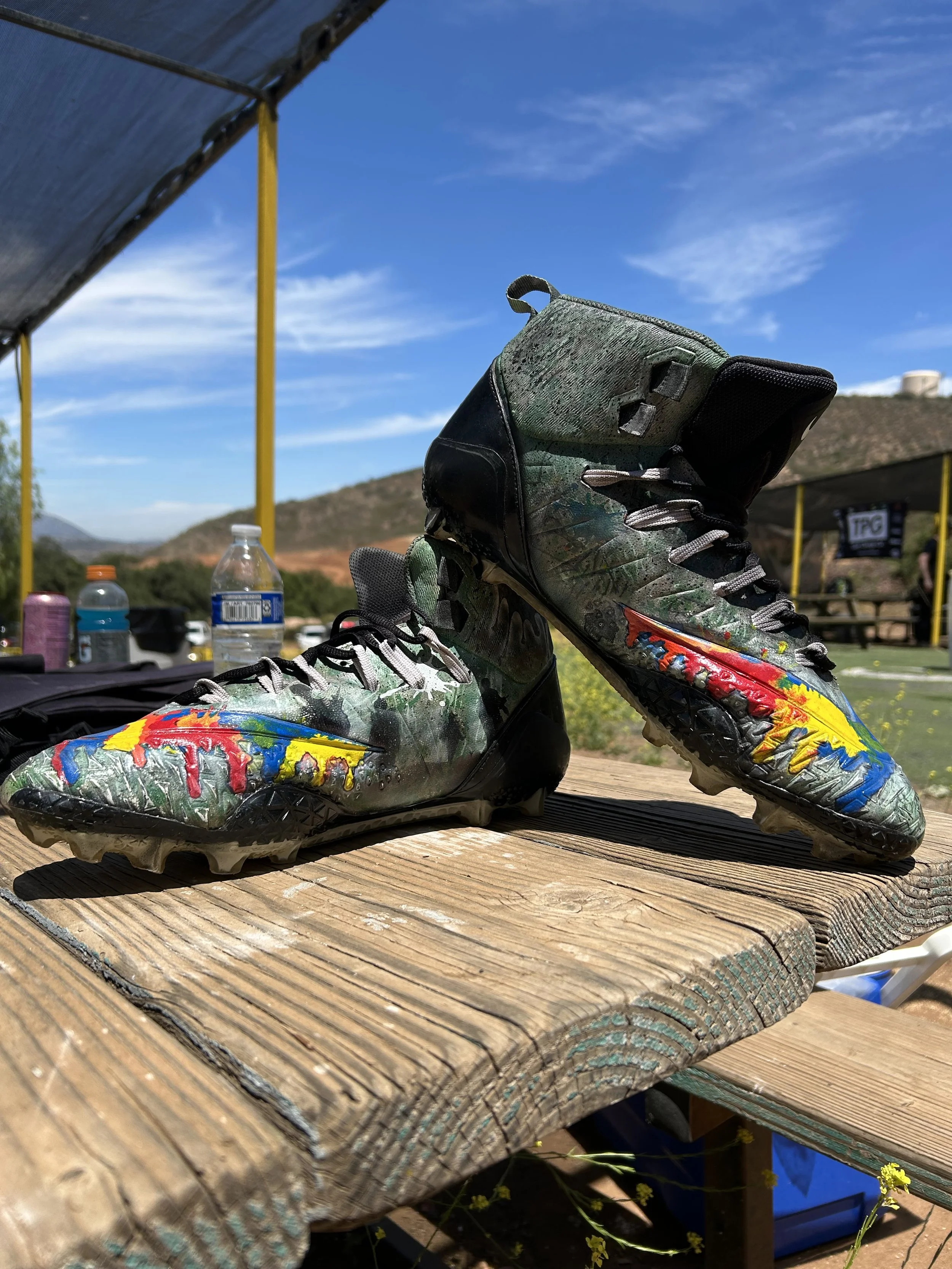A pair of colorful football cleats with a paint splatter design resting on a wooden table outdoors under a canopy, with a background view of a blue sky, hills, and a tent.