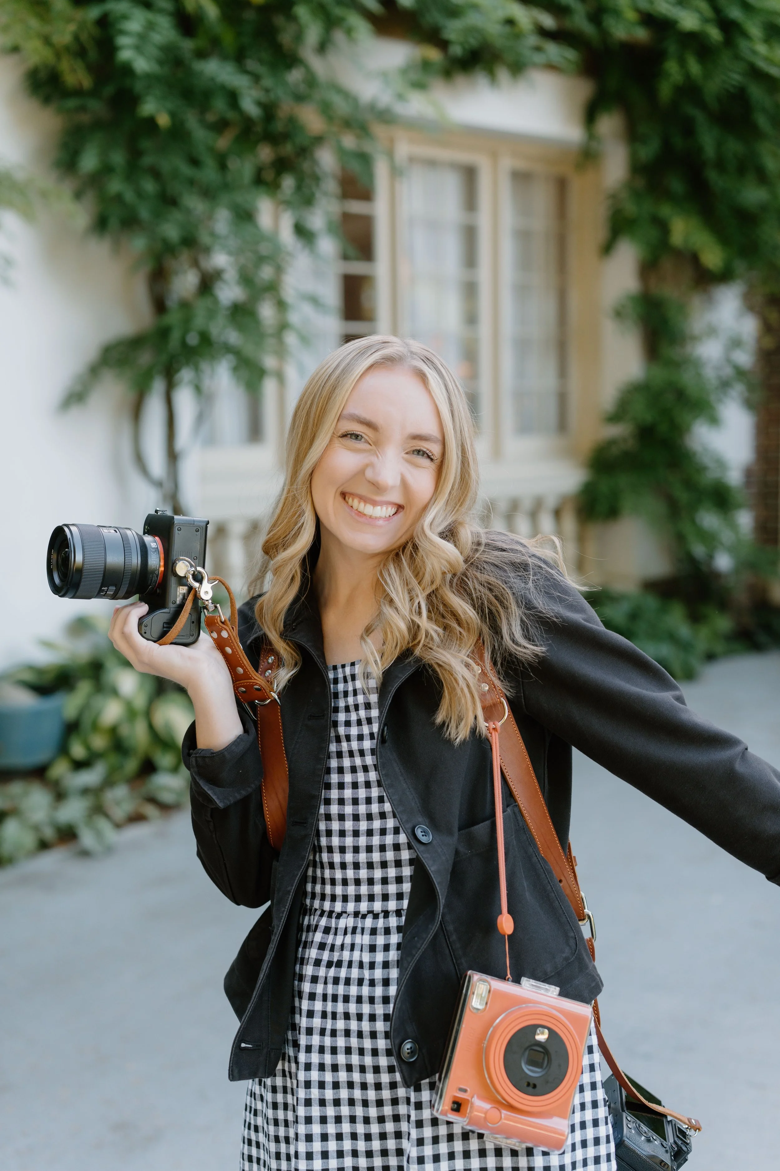 Photo of a wedding photographer wearing a dual harness camera strap, orange Instax camera, and gingham dress, smiling big at the camera while she poses for her headshots at Lairmont Manor, a beautiful and historic wedding venue in Bellingham, WA