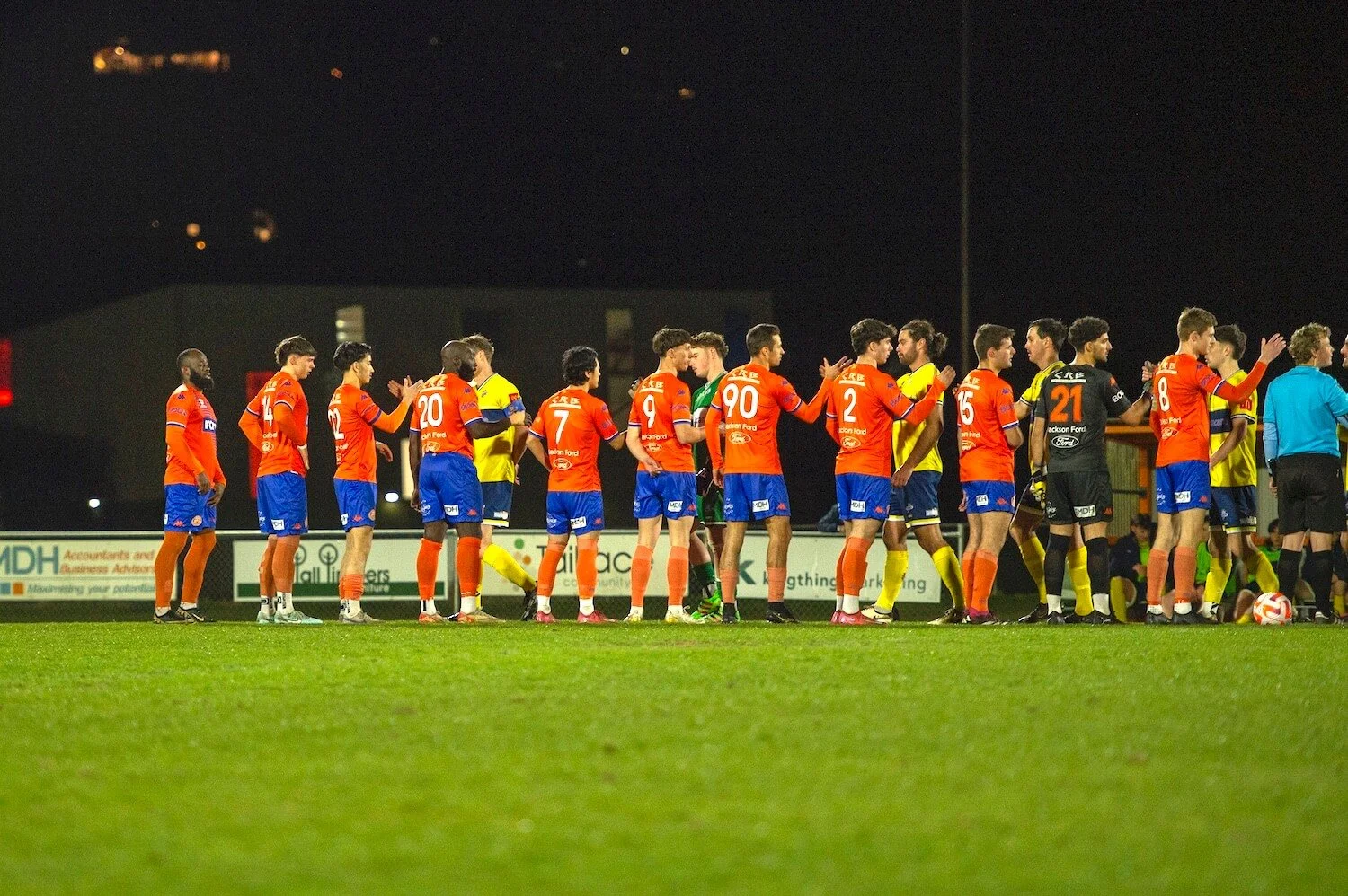 Soccer players in orange and blue uniforms exchanging handshakes on a field at night after a match.