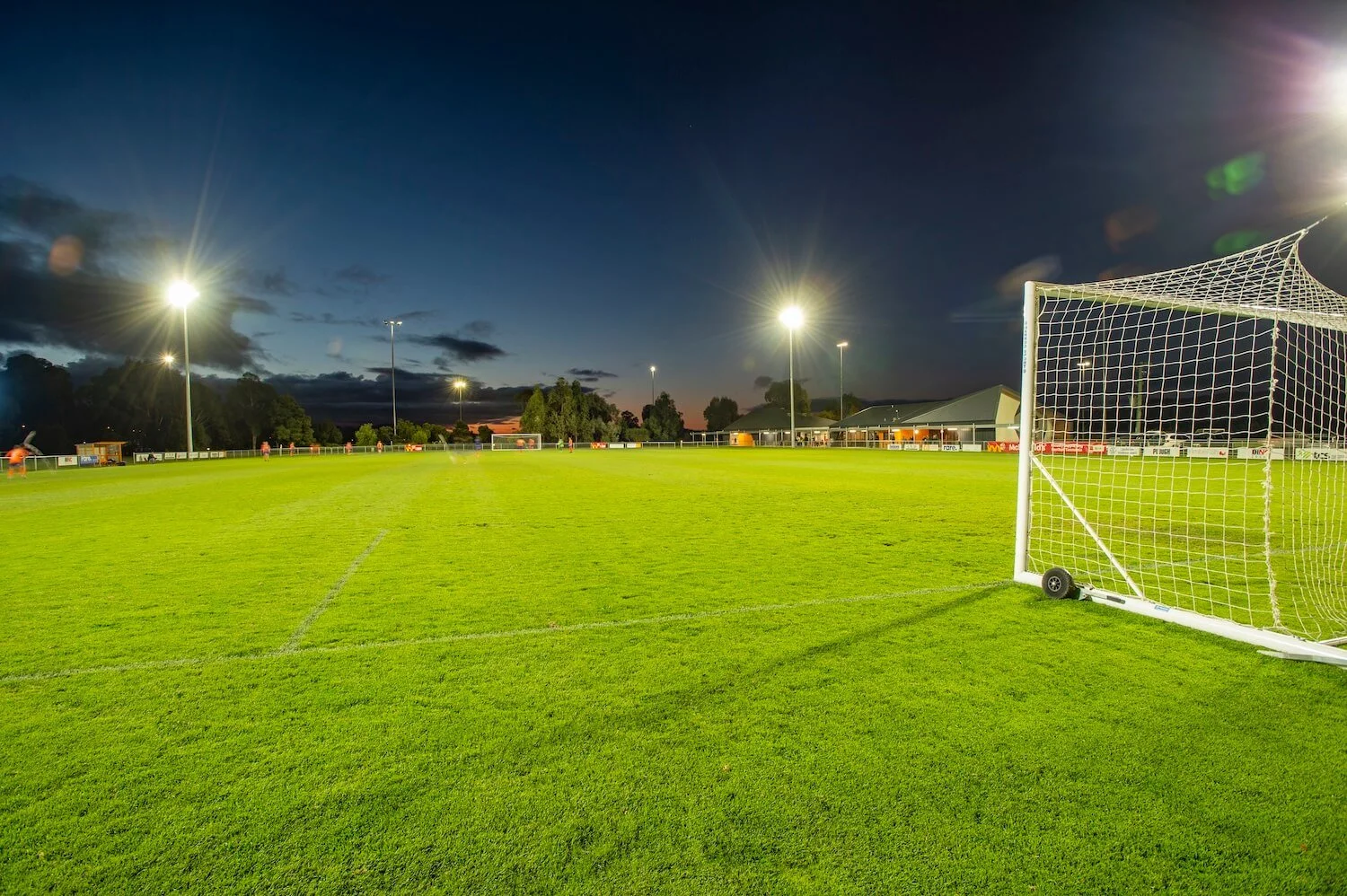 A soccer field during twilight with bright floodlights, a goalpost in the foreground, and players practicing in the background.