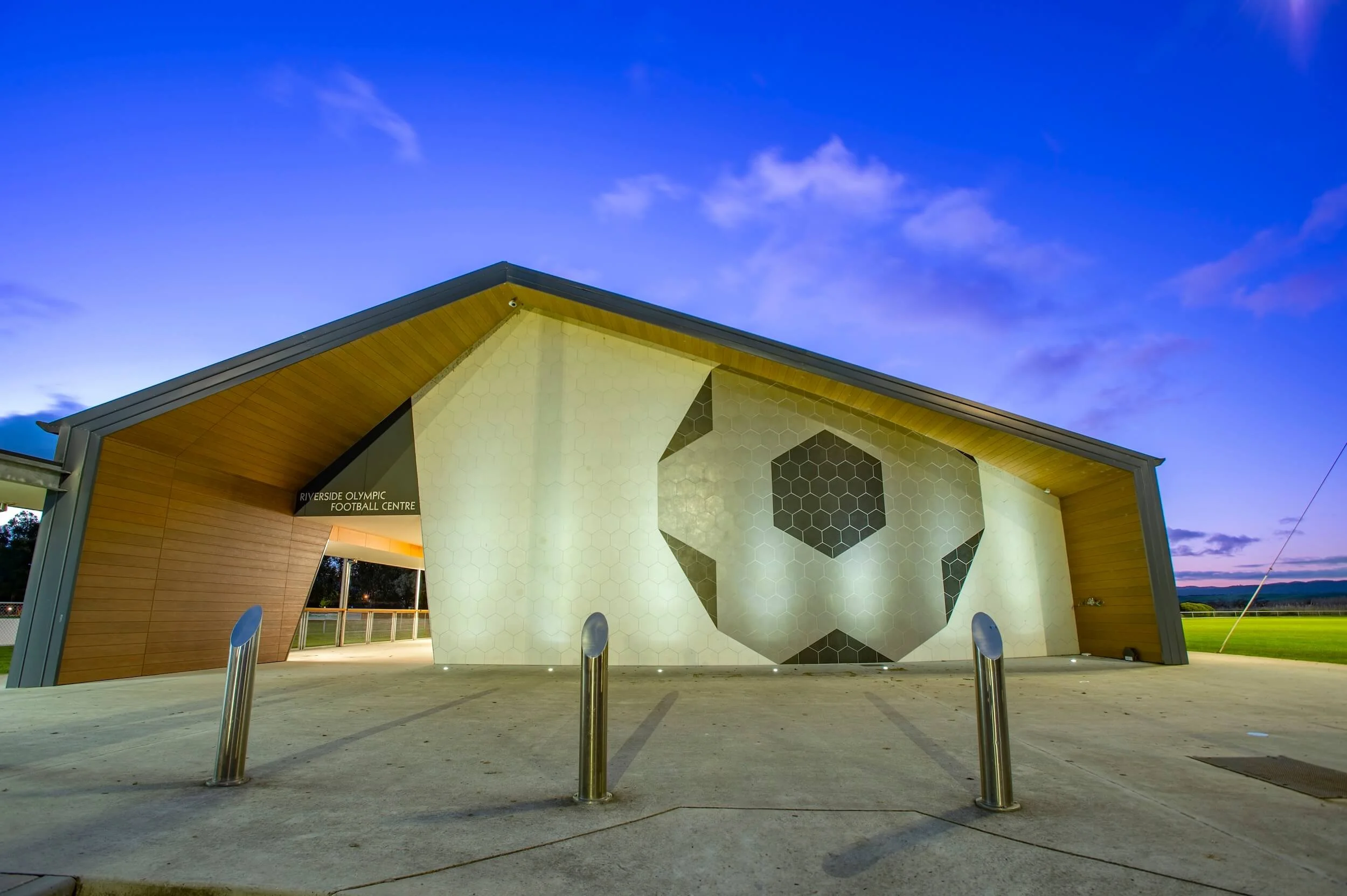 Riverside Olympic Football Centre building exterior at dusk, with a large soccer ball graphic on the wall and a blue sky in background.