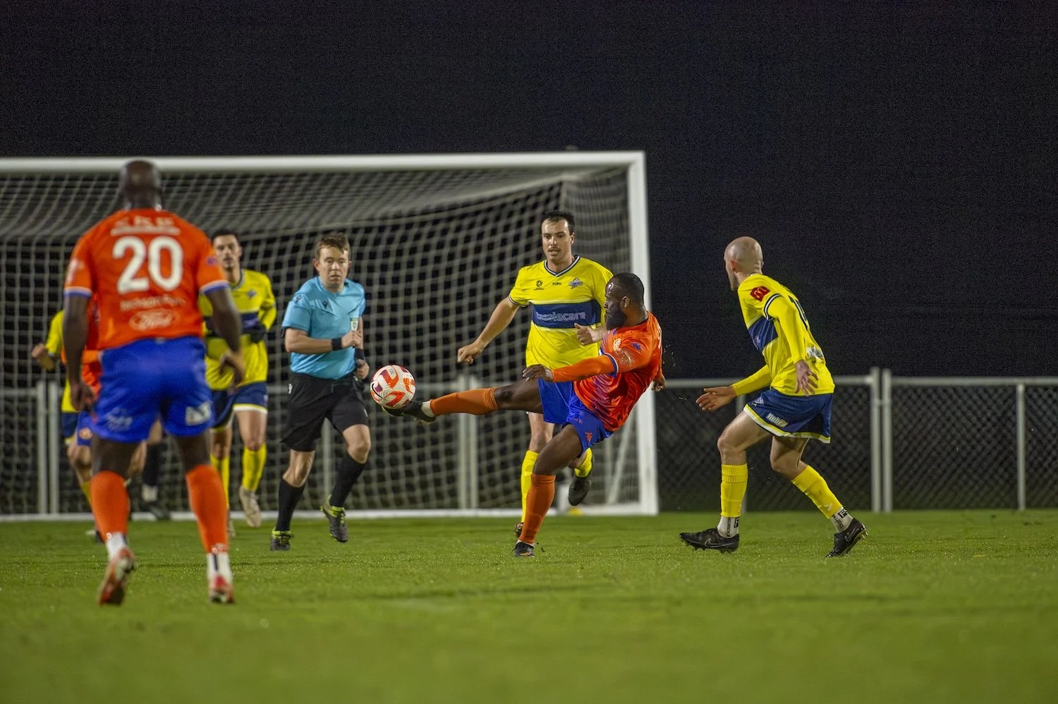 Soccer players in orange and yellow uniforms compete for the ball near the goal during a match under floodlights at night.