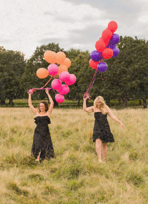 Two women in black dresses holding colorful balloons in a grassy field with trees in the background.