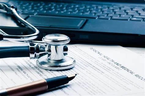 Stethoscope, pen, and medical records on a desk with a laptop in the background.