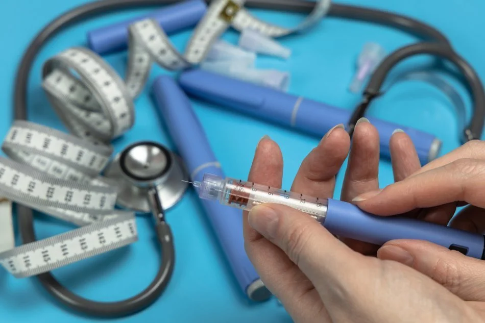 A hand holding a syringe with a blue pen-style handle, surrounded by medical tools including measuring tapes, a stethoscope, and blue medical devices on a blue background.