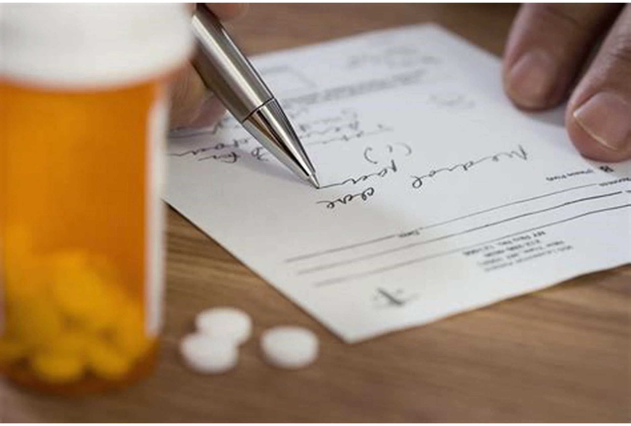 A person writing a handwritten note with a silver pen on a small piece of paper on a wooden table, with an orange pill bottle and white pills nearby.