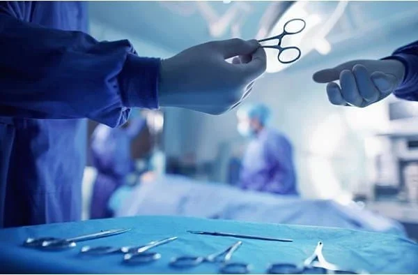 Close-up of a surgeon handing surgical scissors to another person in an operating room, with surgical tools on a table in the foreground and medical staff in surgical gowns and masks in the background.