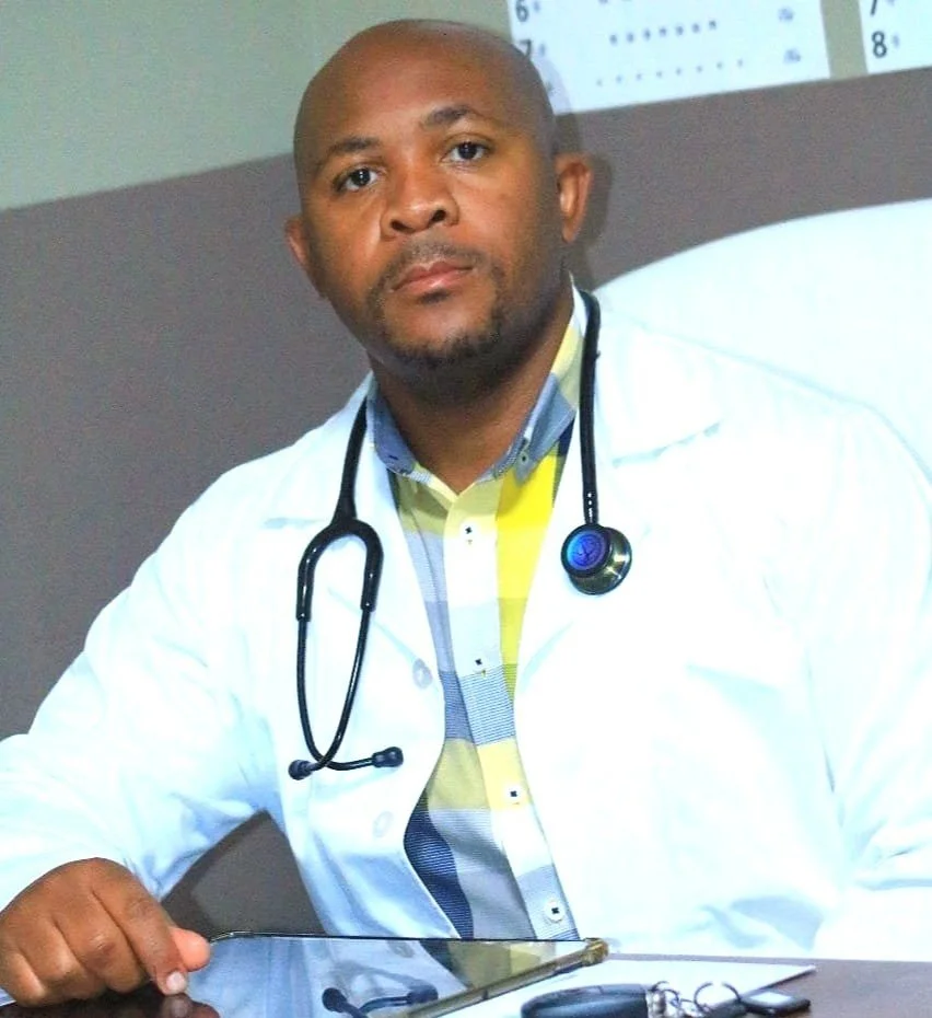 A male doctor wearing a white coat with a stethoscope around his neck, sitting at a desk with a tablet and car keys.