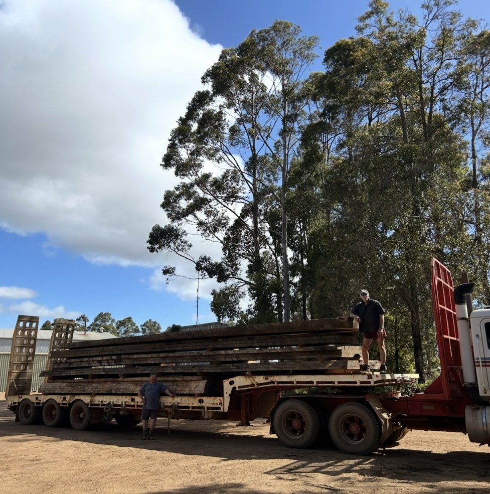 Two men standing next to a loaded flatbed truck carrying lumber in an outdoor area with tall trees and a partly cloudy sky.