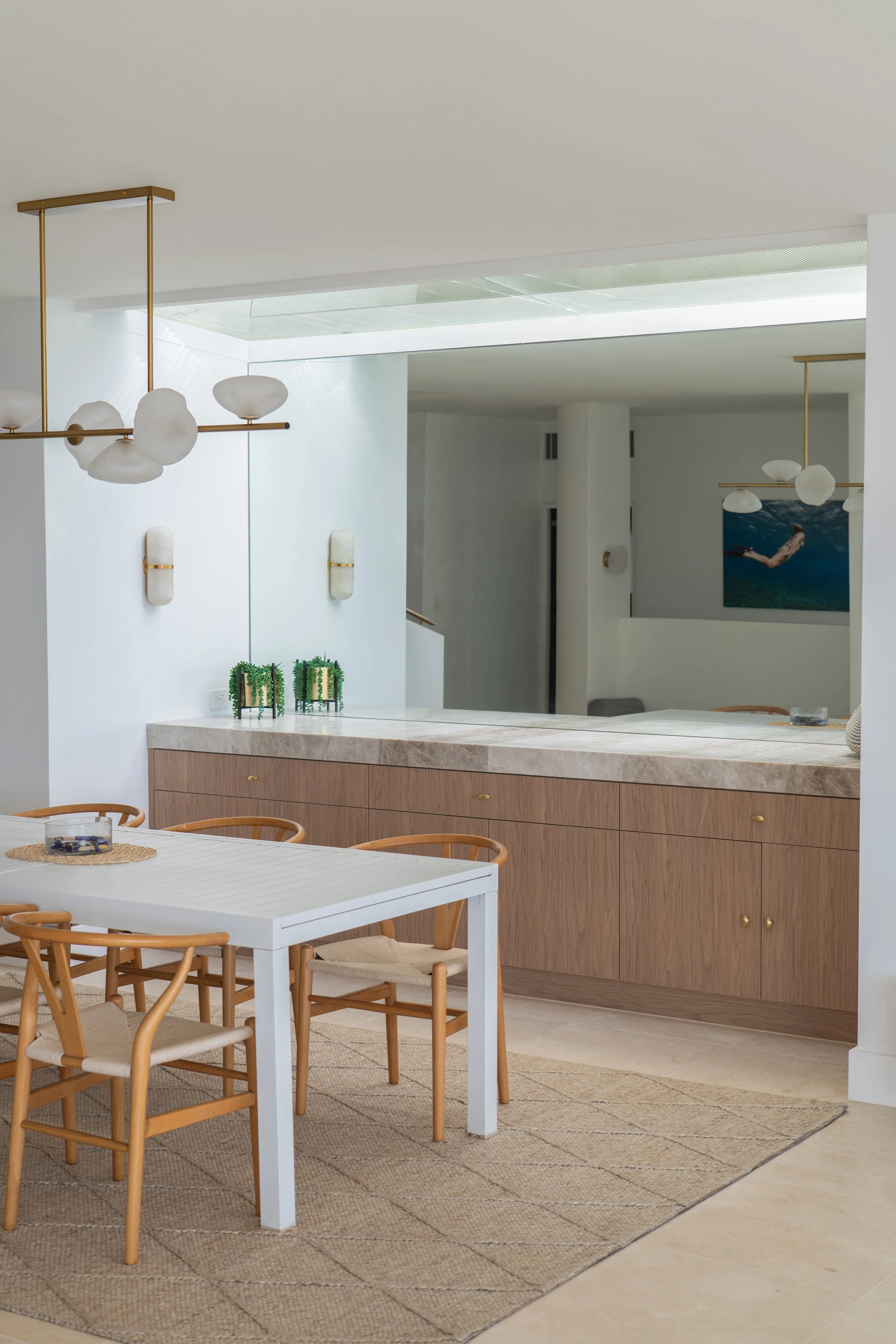 Modern dining area with a white table, wooden chairs, and a beige rug. The background features a light wood cabinet, white walls, and hanging light fixtures with white globes and brass accents.