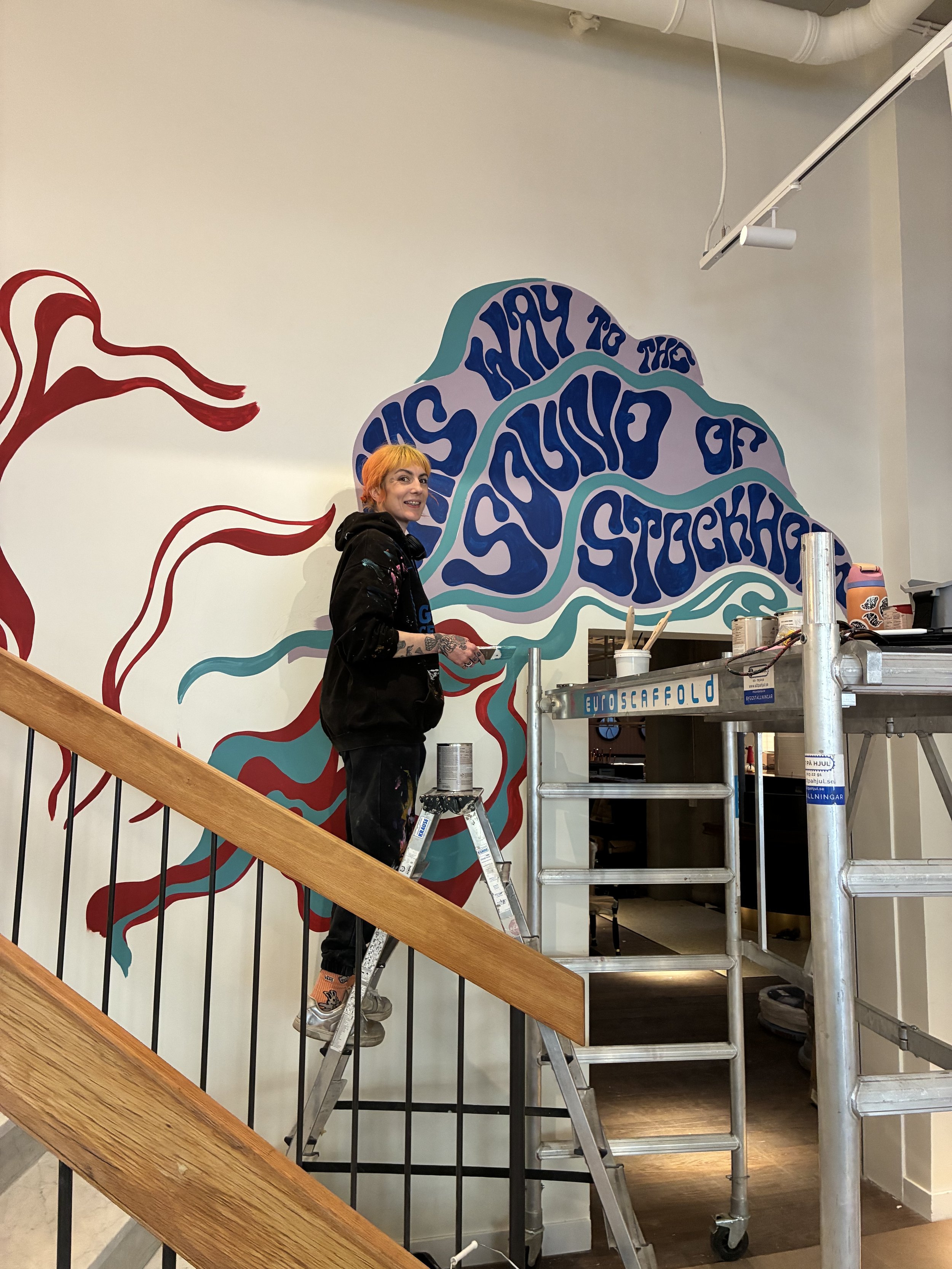 hand lettering artist illustrator and muralist thirteen scorpios stands on a ladder painting a 15ft mural