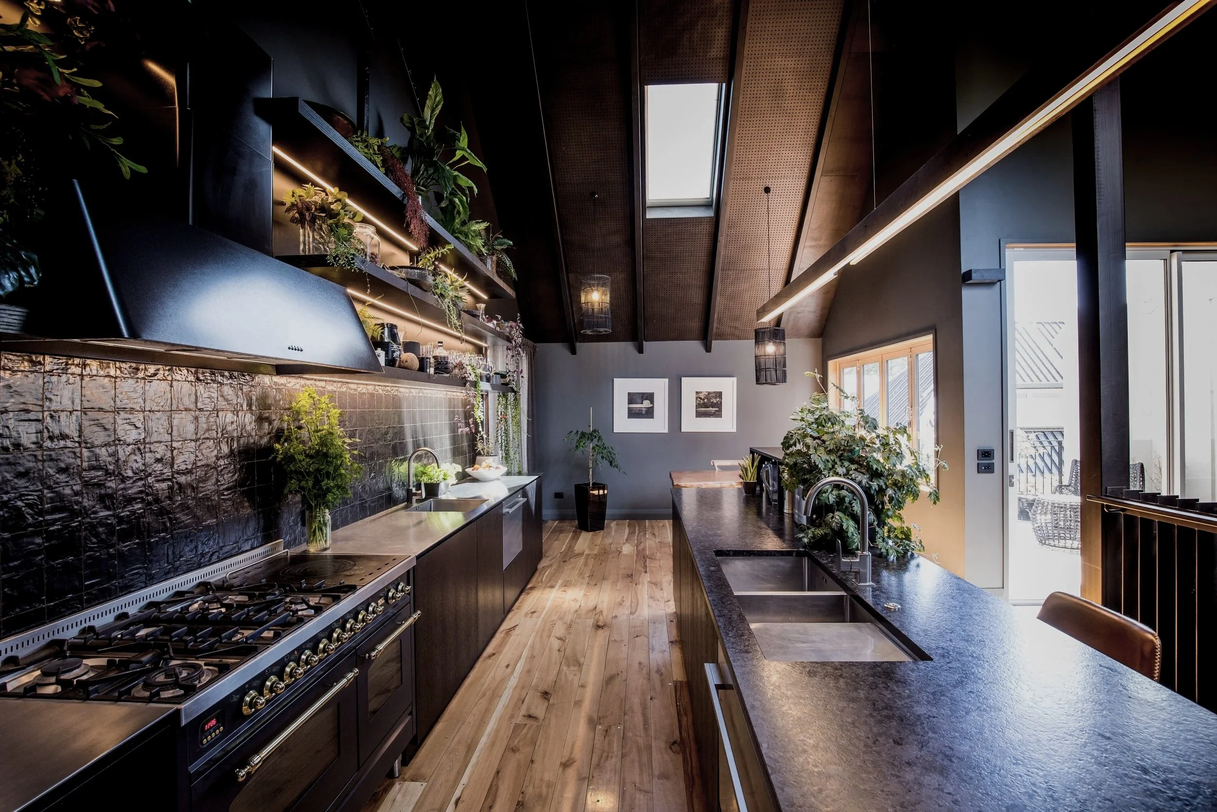 Interior design by The Nave, Jo Grams Modern kitchen with black cabinets, a large gas stove, black tile backsplash, and wooden floors. Shelves with plants and decor under a sloped ceiling with skylight, and a sliding door leading outside.