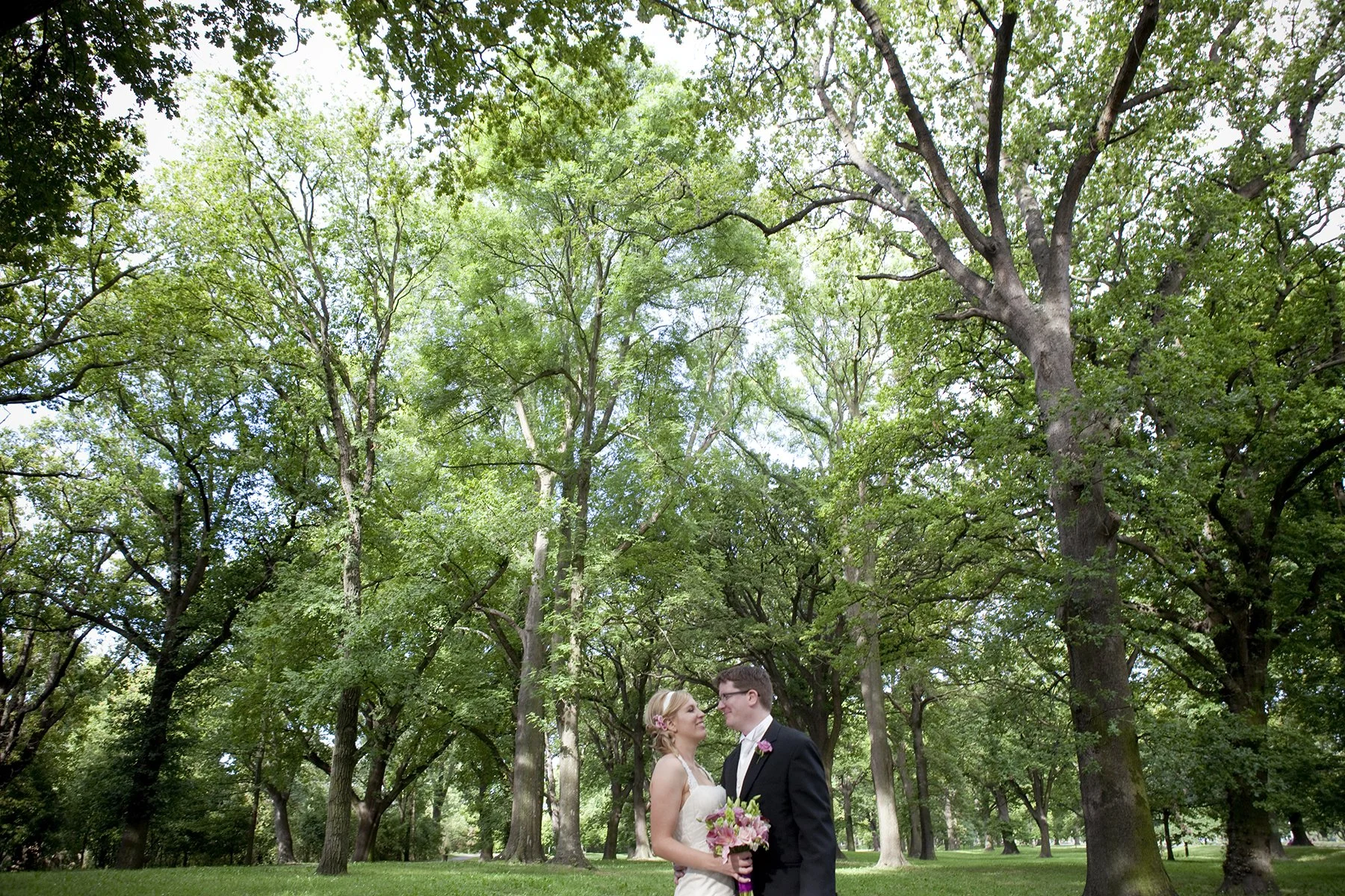 A bride and groom standing close together in a park with tall trees and green foliage, looking into each other's eyes during their wedding photos.
