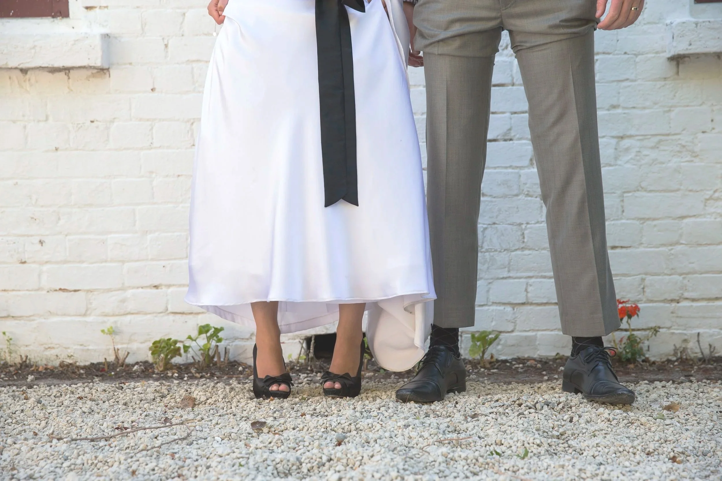 Close-up of a bride and groom's lower bodies, showing the bride in a white gown with black high heels and the groom in gray suit pants with black shoes, standing against a white brick wall.