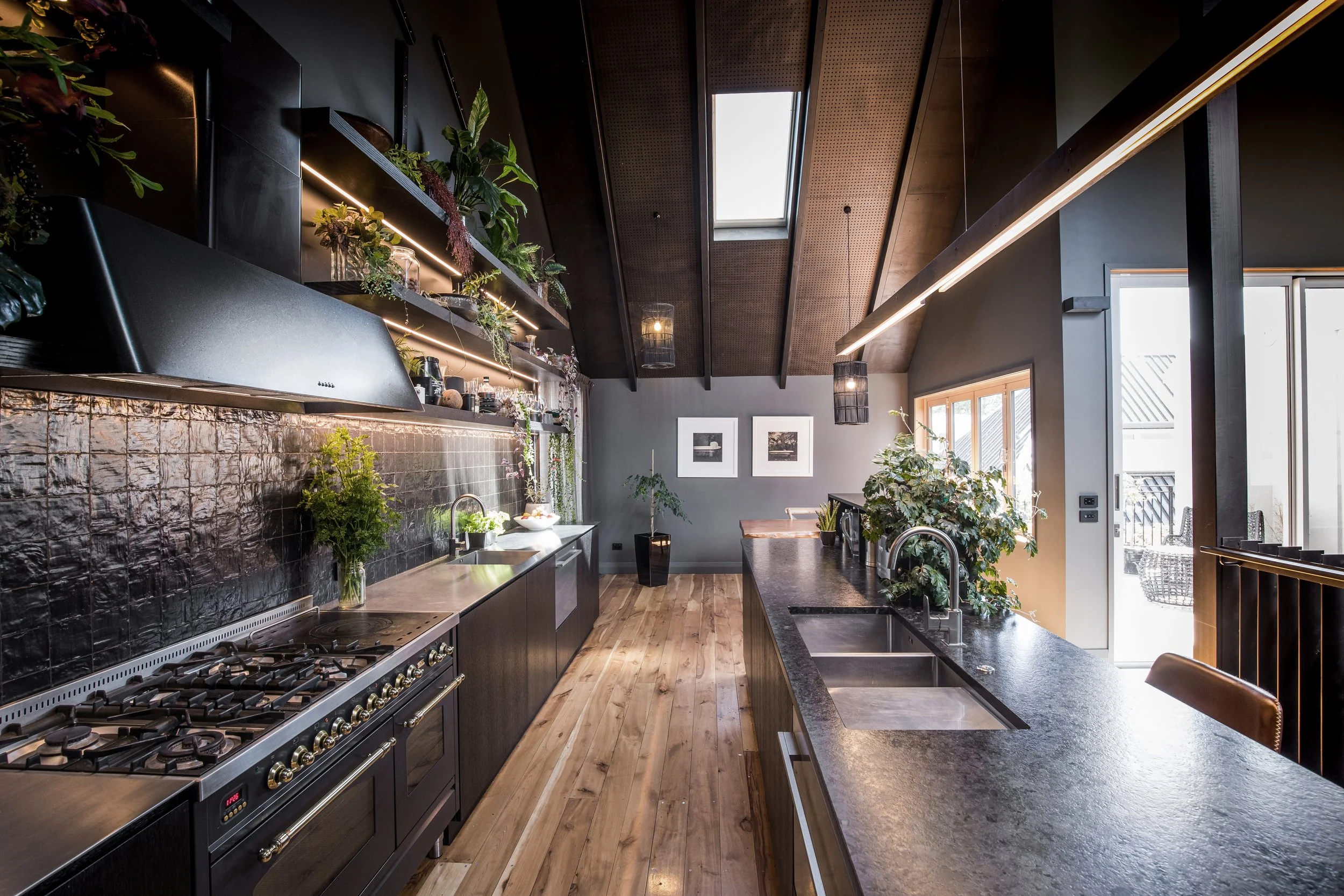 Modern kitchen with black cabinets, a large gas stove, and black countertops. Open shelving with plants and decorative items. Wooden floors, a skylight, and large windows letting in natural light. Dining area with plants and framed pictures on the wall.