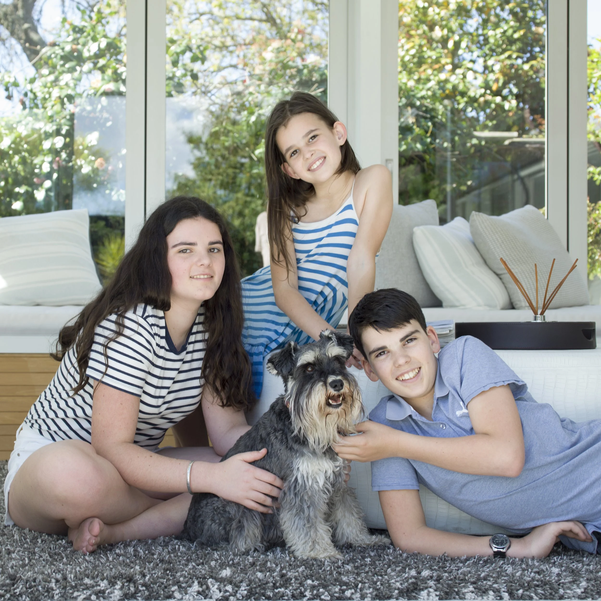 Four children and a dog inside a bright living room with large windows and greenery outside.