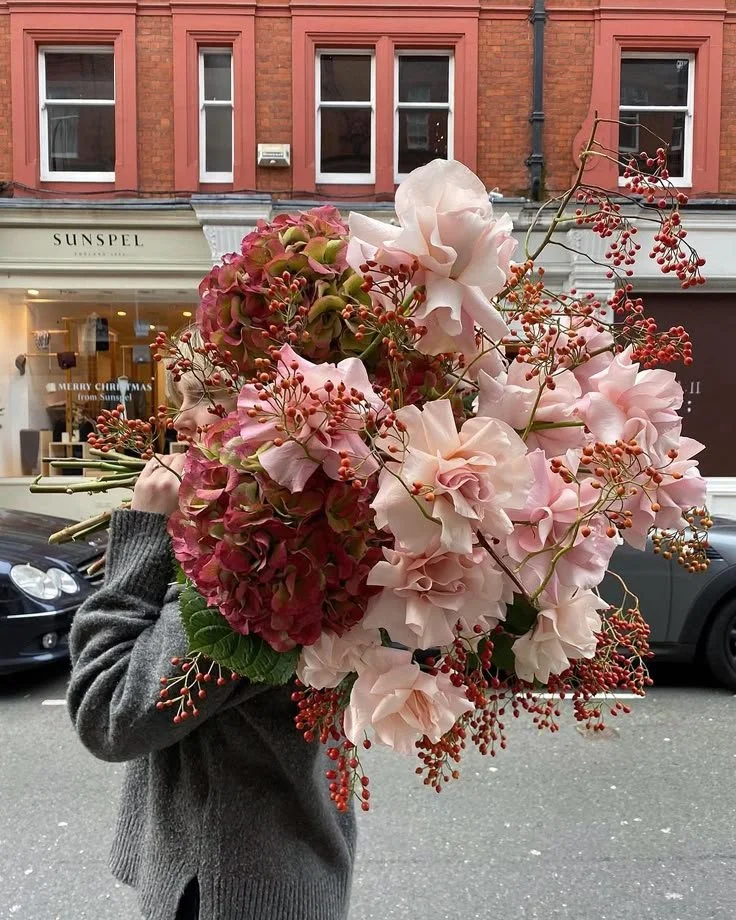 A person holding a large bouquet of hydrangeas, roses, and berries on a city street with storefronts and cars in the background.