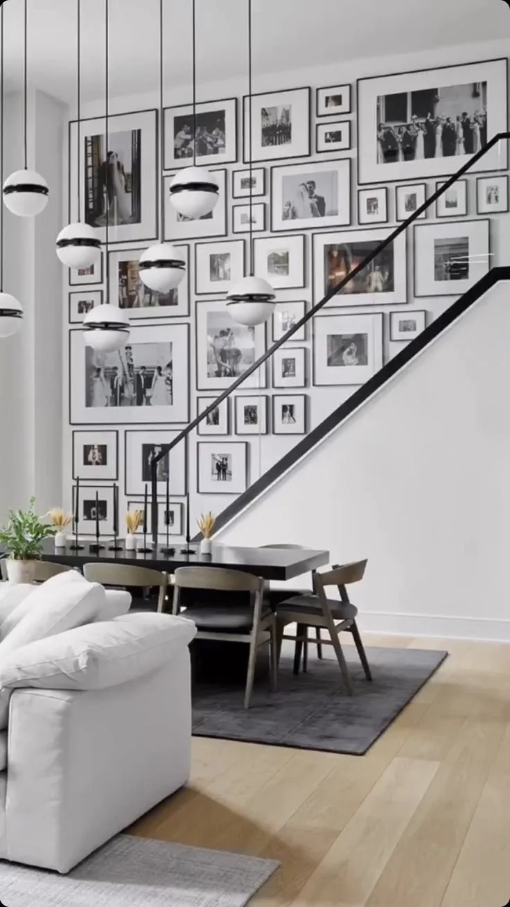 A modern living room and dining area with a gallery wall of black-and-white photographs behind a staircase. The space features hanging pendant lights, a white sofa, and a dark dining table with matching chairs. There is a small rug under the dining table and some potted plants on the side.