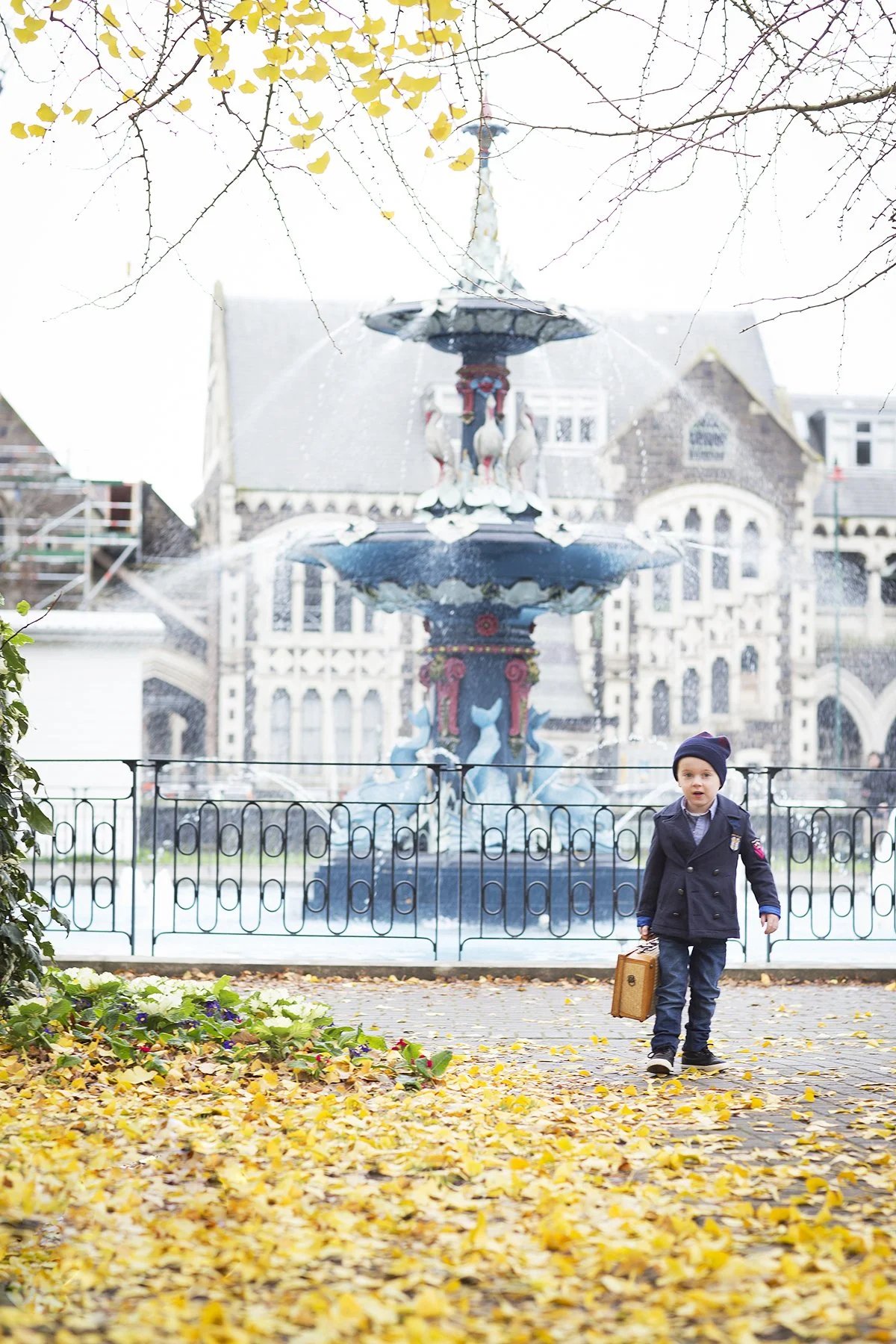 A young boy walking on a paved path covered in fallen autumn leaves, holding a small suitcase, with a vintage-style fountain featuring dolphins and a house with ornate architecture in the background.
