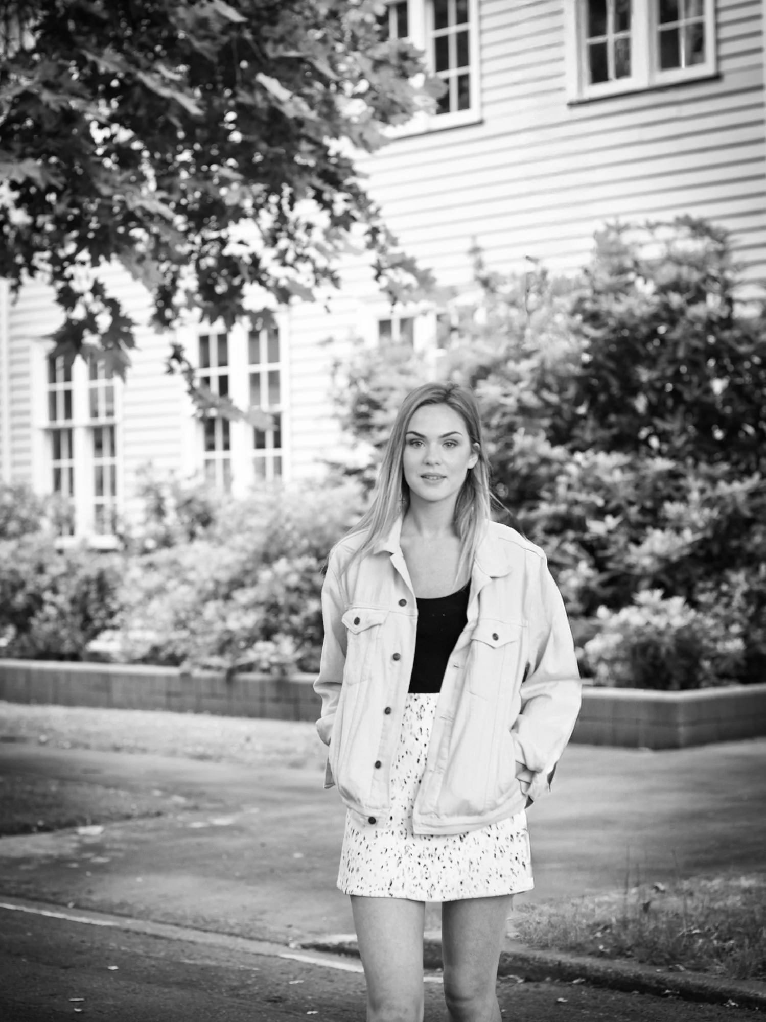 A young woman standing outdoors on a paved surface with a house and trees in the background, wearing a light-colored denim jacket, black top, and a patterned skirt, looking at the camera.