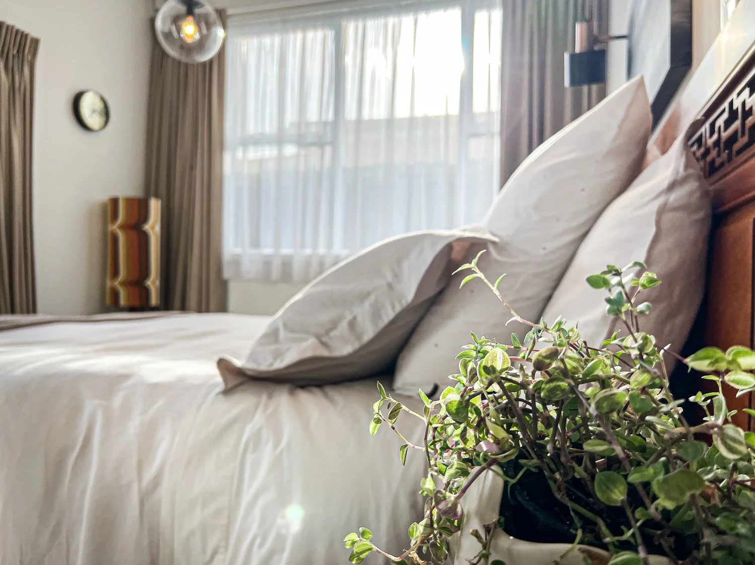 A neatly made bed with white sheets and pillows in a bedroom with a window, curtains, and a potted green plant in the foreground.