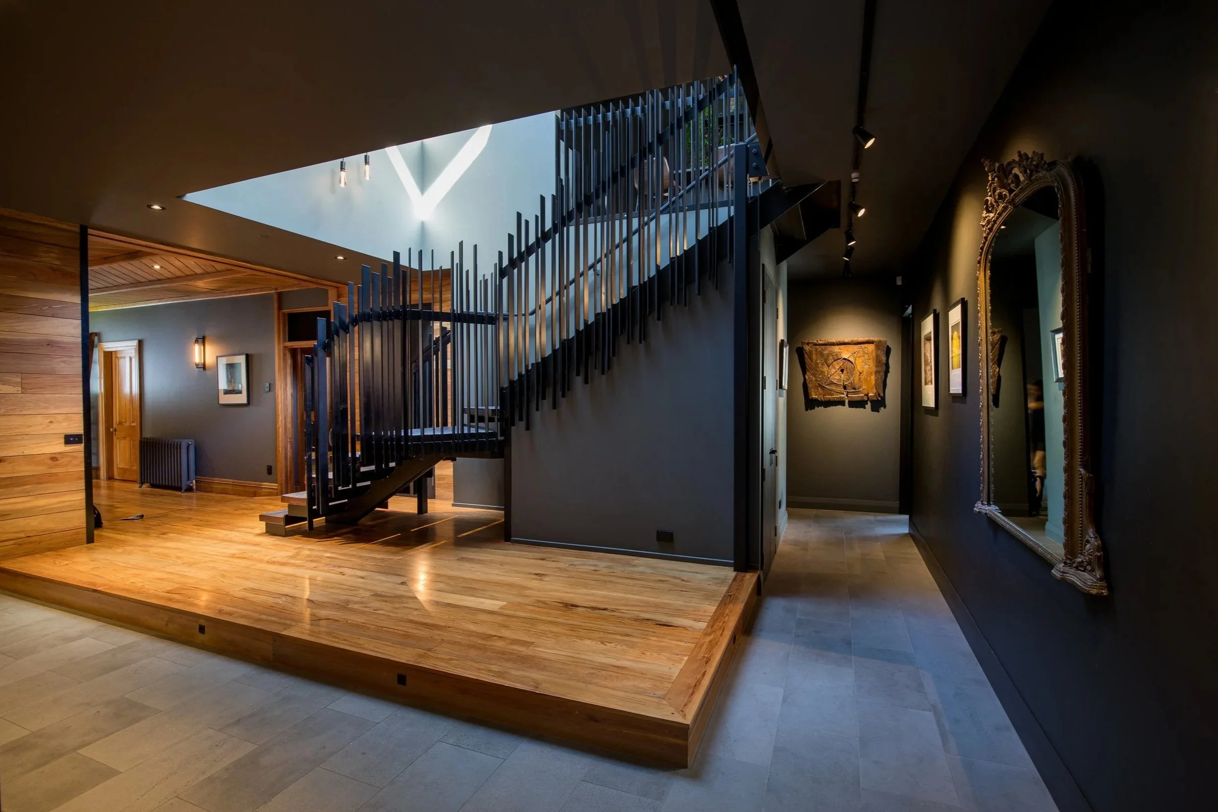 Interior of a house with a staircase, wooden flooring, black walls, large ornate mirror, and artwork on the walls.