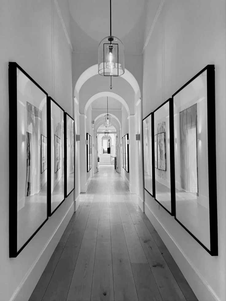 A long hallway with arched ceilings, hanging pendant lights, and framed artwork on both sides, leading to a bright doorway at the end.