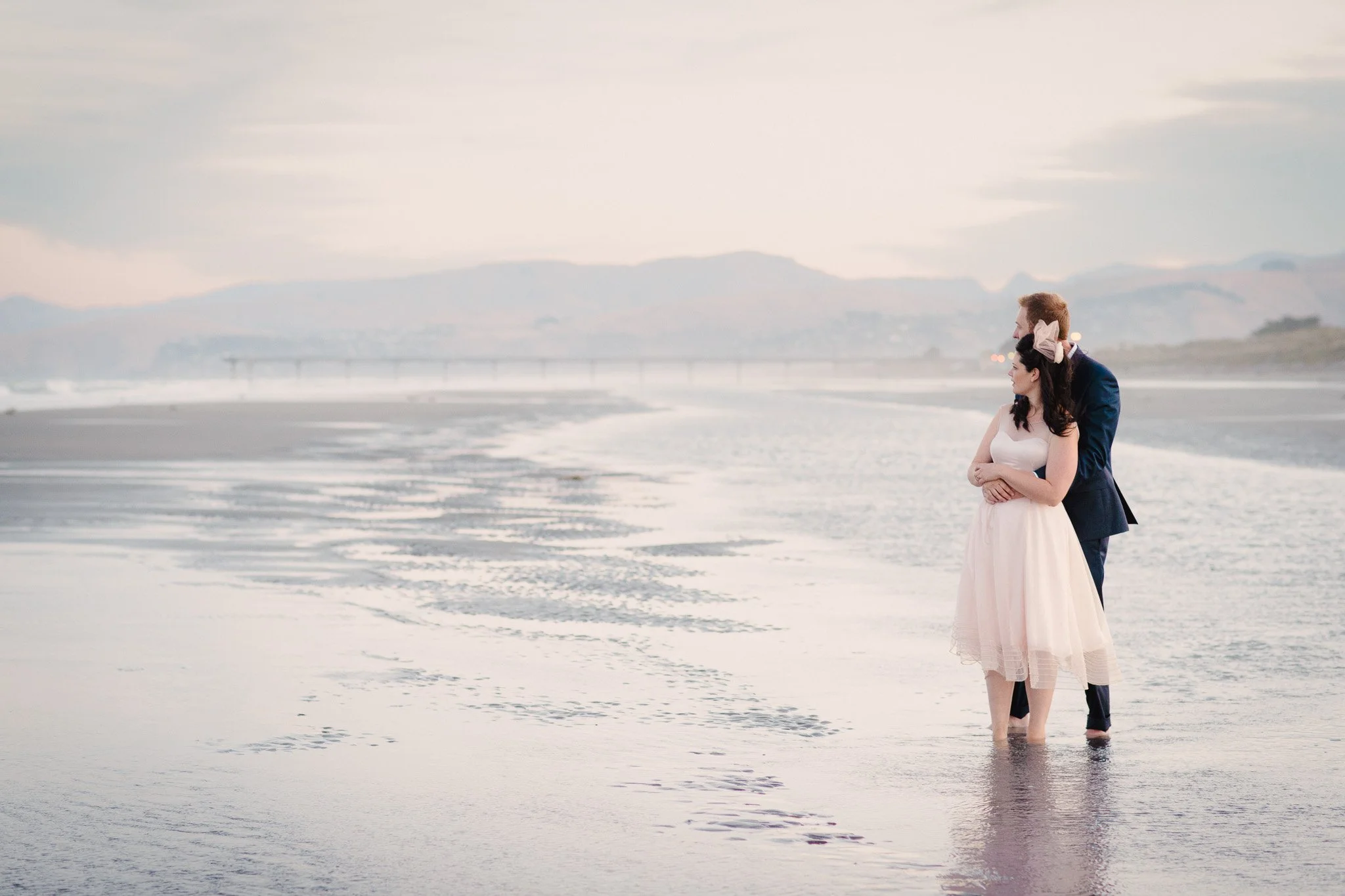 A couple standing in the shallow water at the beach, embracing and looking into the distance, with mountains and a bridge in the background during sunset.