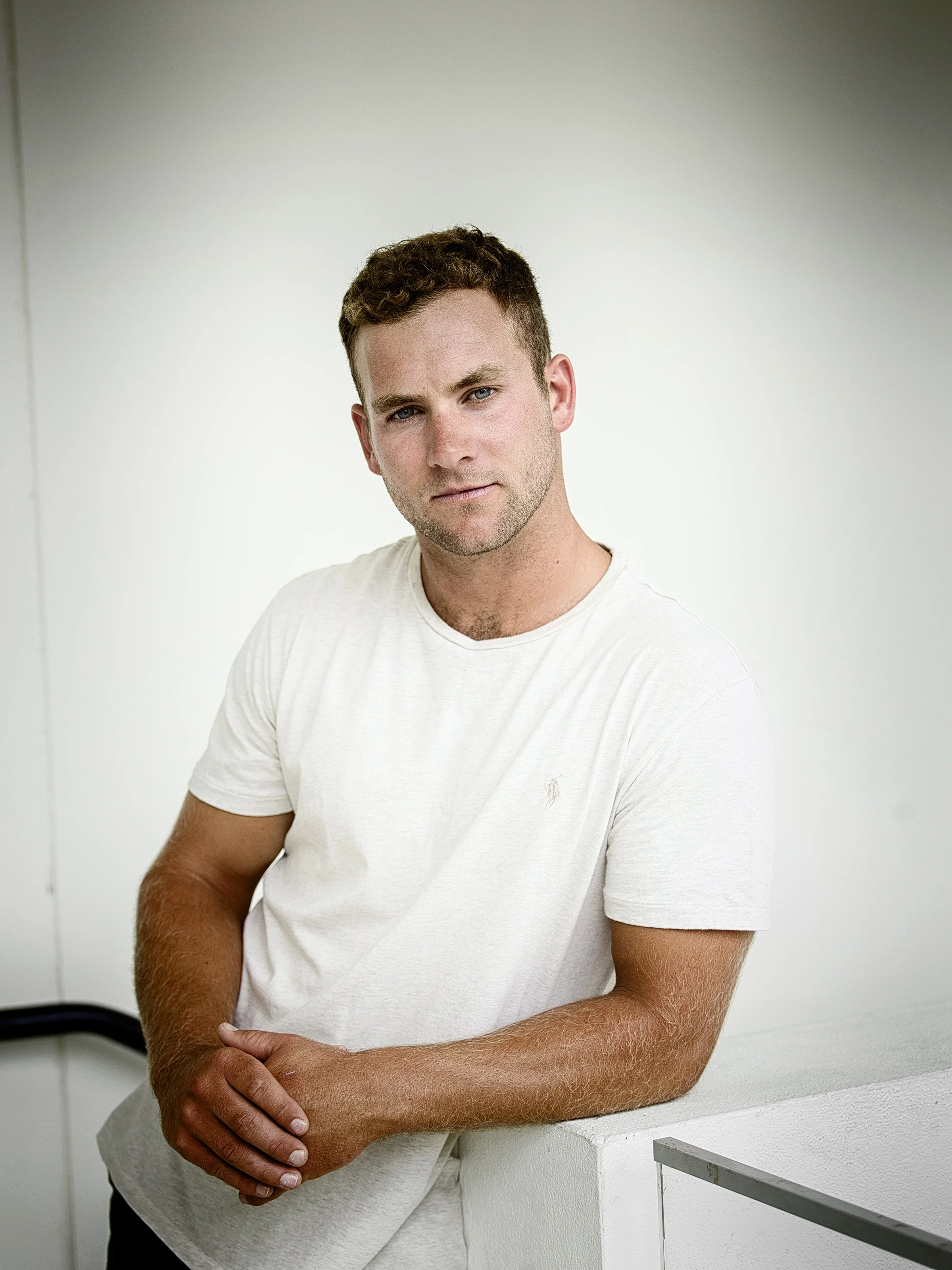 Young man with short brown hair and fair skin leaning on a white surface, wearing a white t-shirt, with a serious expression, against a plain light-colored background.