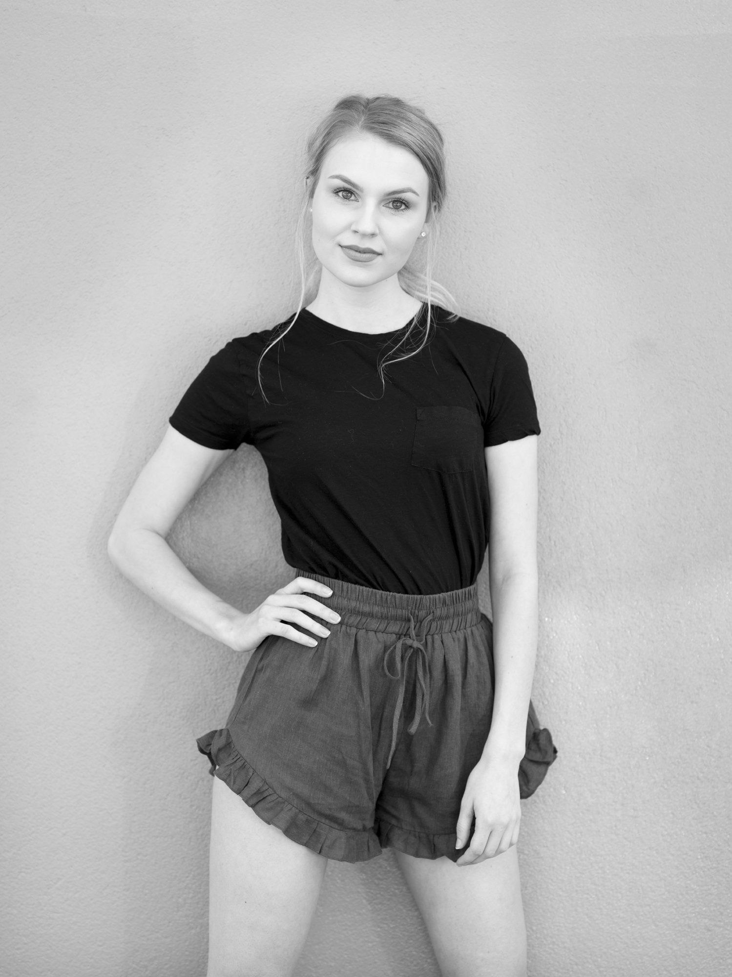 A young woman with light hair styled in a loose ponytail, wearing a black t-shirt and shorts, standing against a plain wall.
