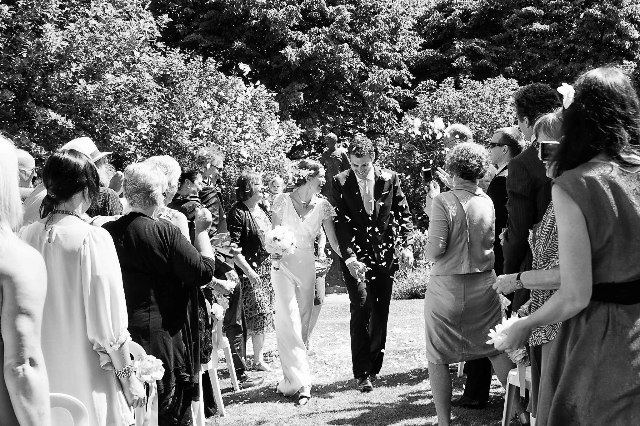 Black and white photo of a bride and groom walking hand in hand down the aisle at an outdoor wedding, surrounded by guests on either side taking pictures and celebrating.