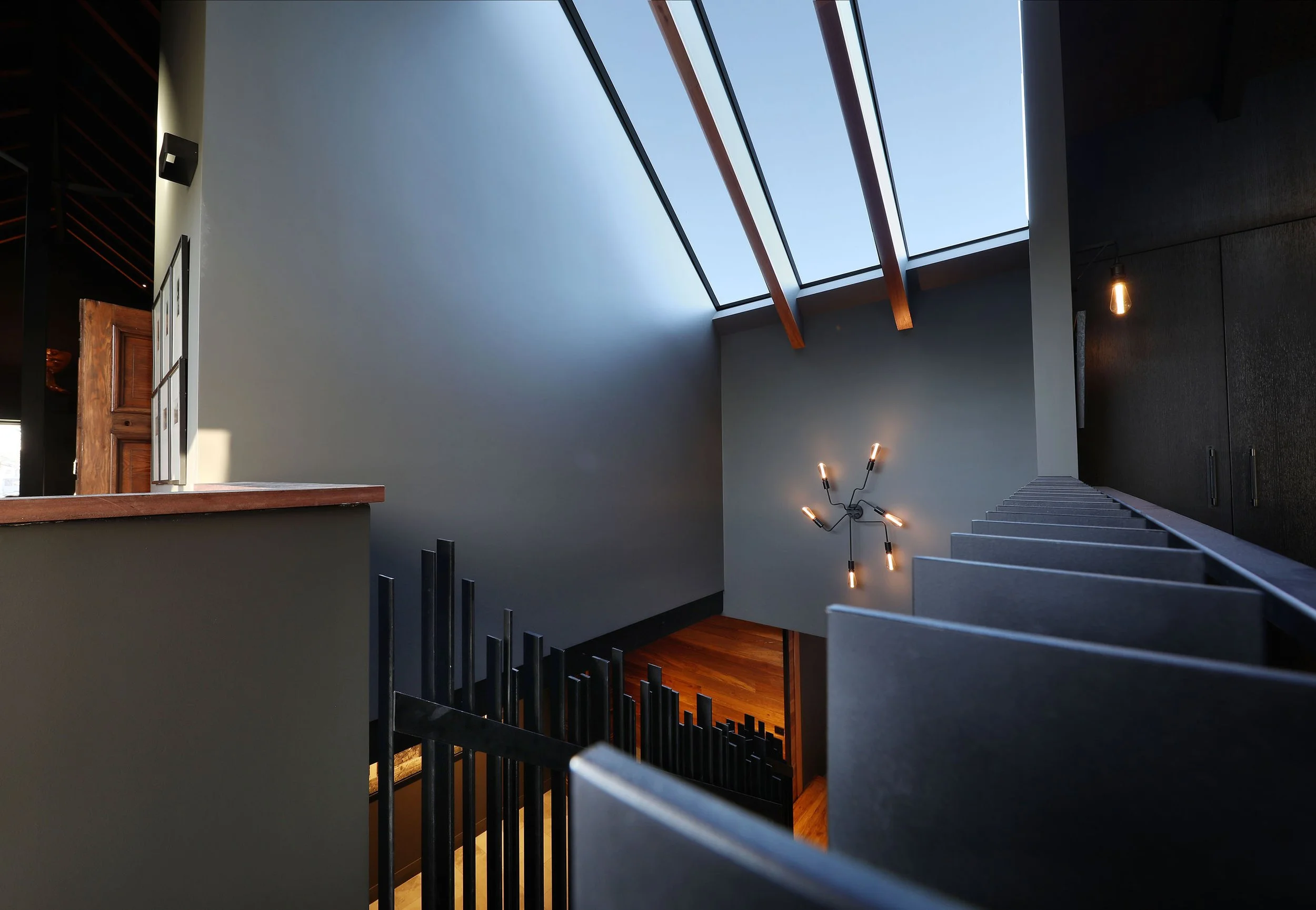 Modern staircase with black metal railing, wooden steps, and a wall with black textured finish. Large skylight above, with blue sky visible, natural light illuminating the space. Contemporary wall-mounted light fixtures with exposed bulbs.