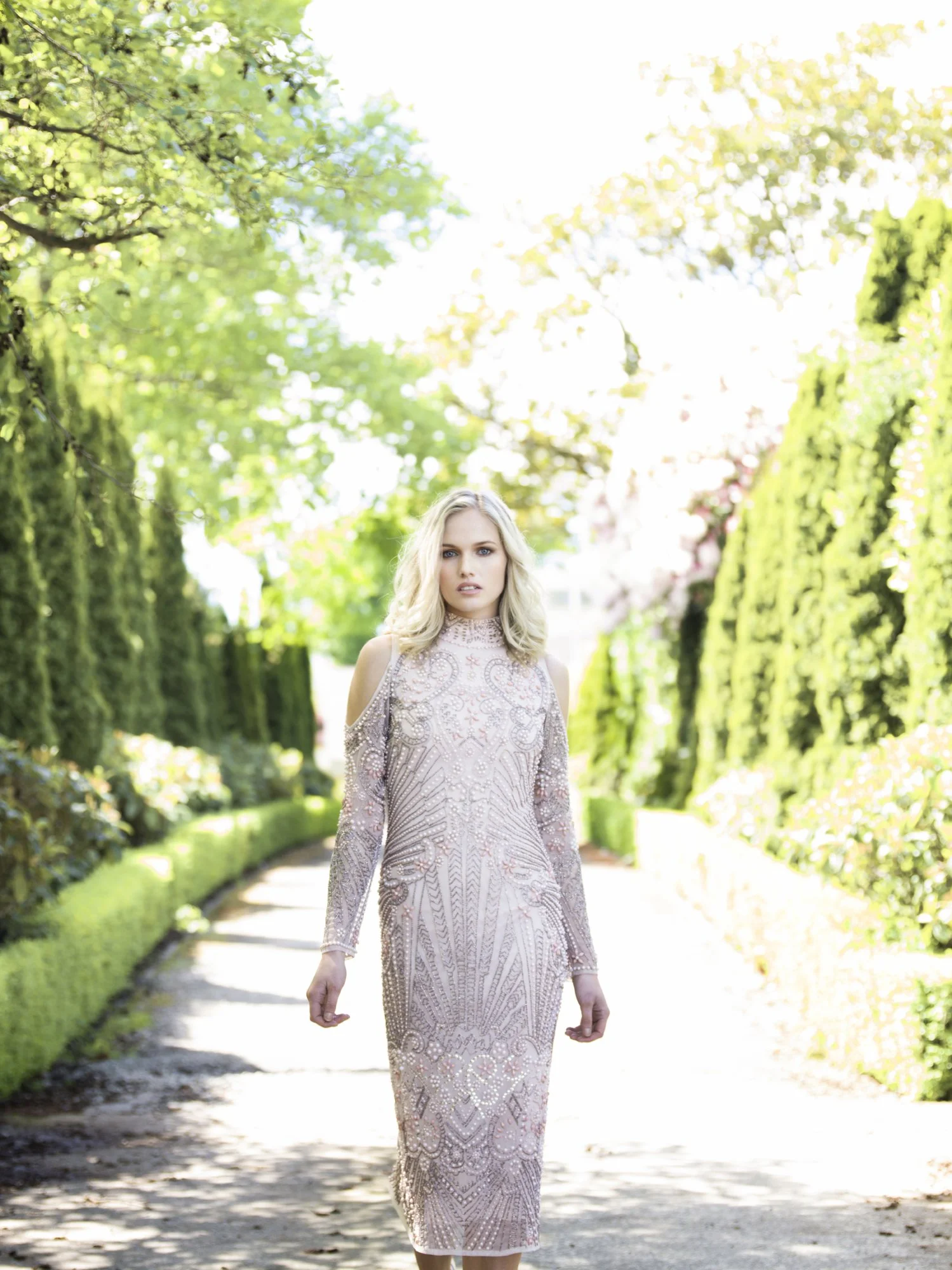 A woman walking in a sunny park or garden, wearing a shimmering, embellished dress with cut-out shoulders and long sleeves, surrounded by green trees and bushes.