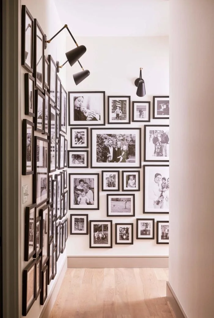 A wall decorated with a gallery of black and white family photos in black frames, illuminated by modern black wall-mounted and ceiling-mounted lamps.