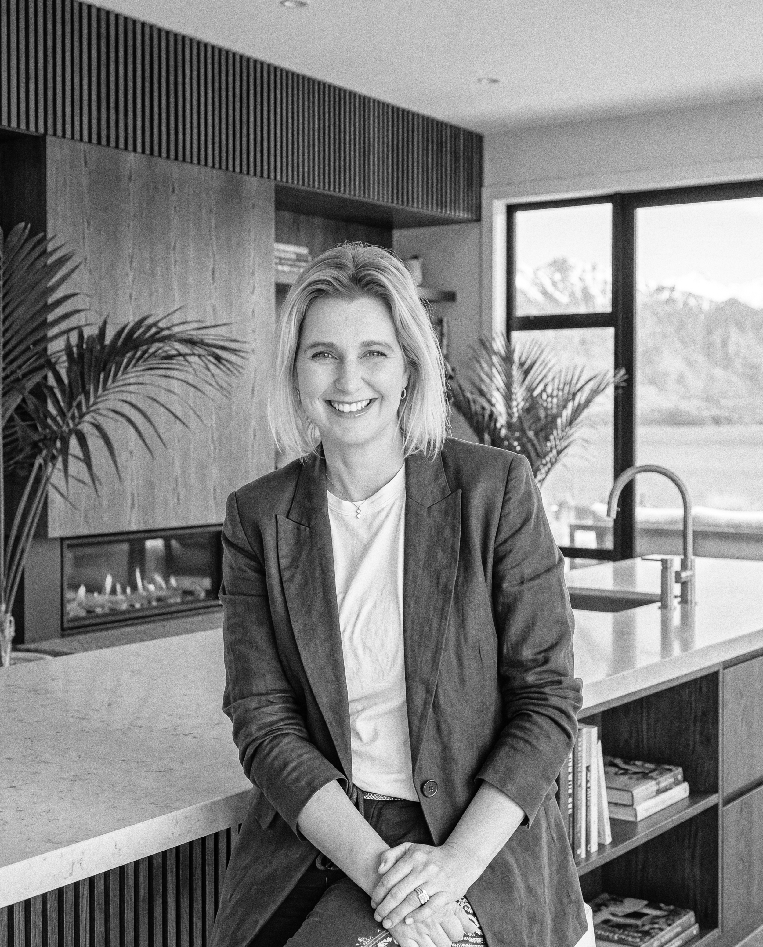 A woman with light hair, wearing a suit jacket and white shirt, smiling and sitting in a modern office or library space with large windows, wooden panels, plants, and books in the background.