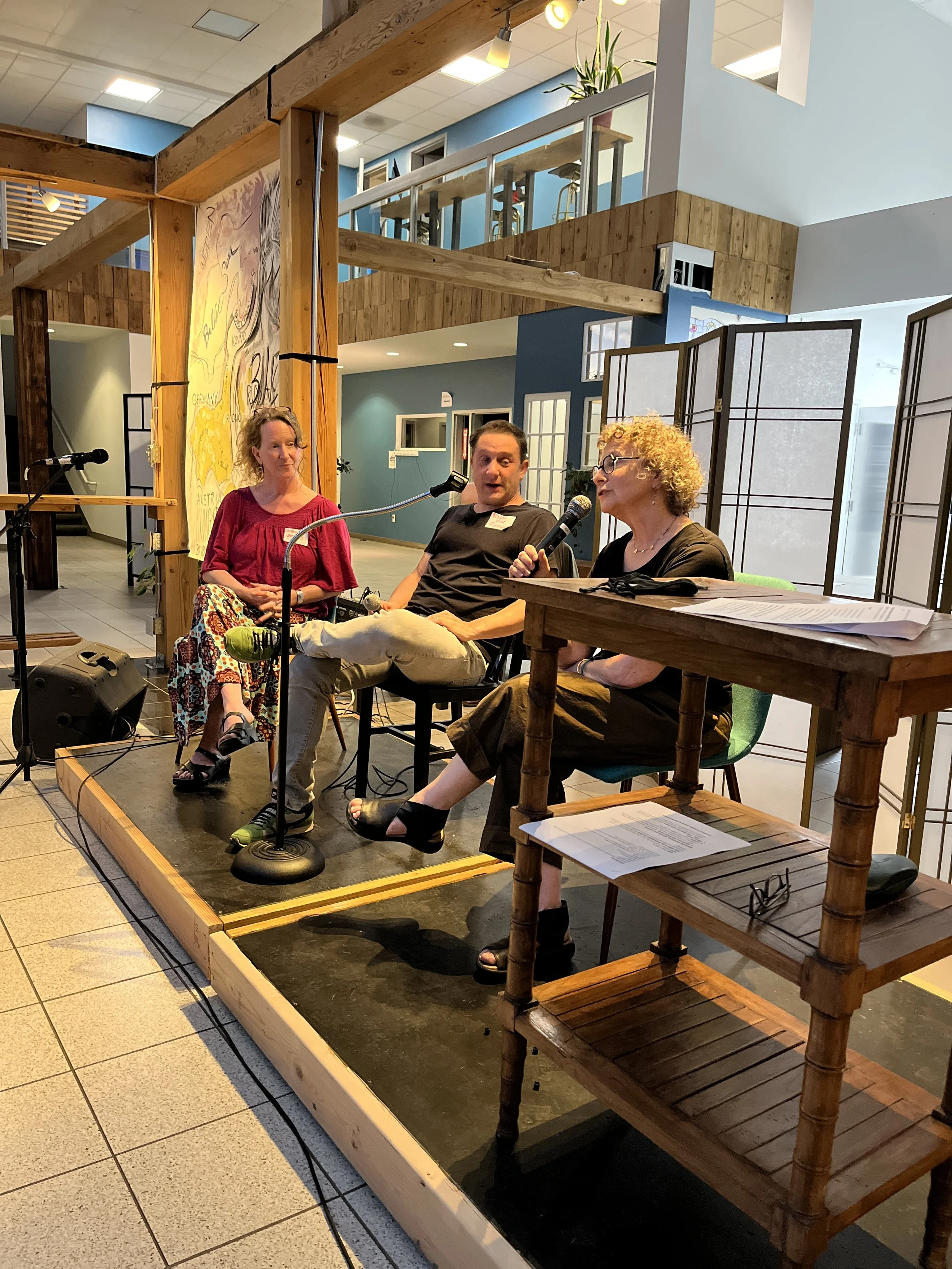 Three people seated on a small stage with microphones, participating in a panel discussion inside a well-lit, modern building with wooden and blue decor.