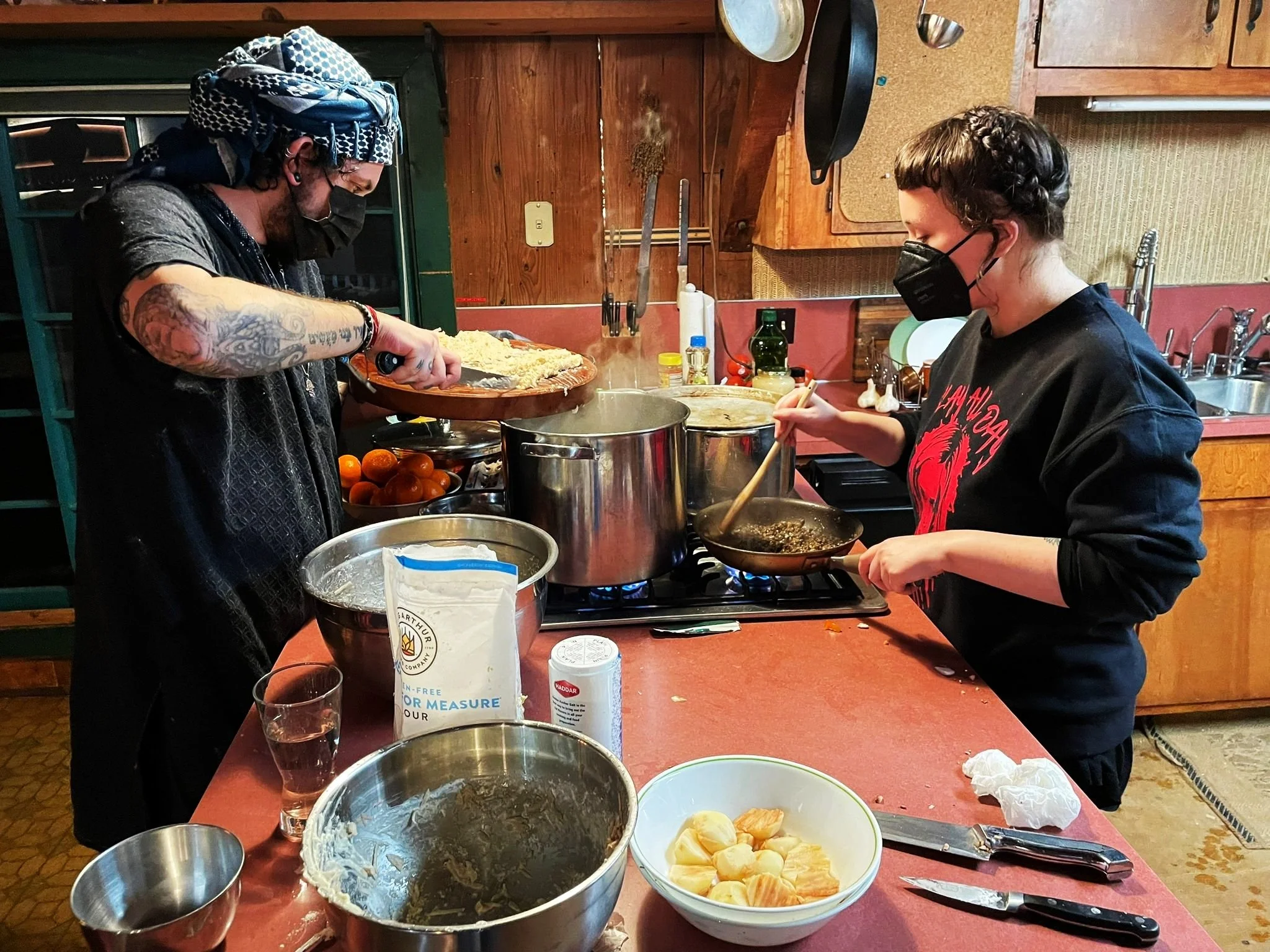 Two people wearing black face masks cooking in a rustic kitchen. The person on the left, with tattoos and a head covering, is scooping food onto a plate. The person on the right is stirring a pan on the stove. The kitchen has wooden cabinets, a count