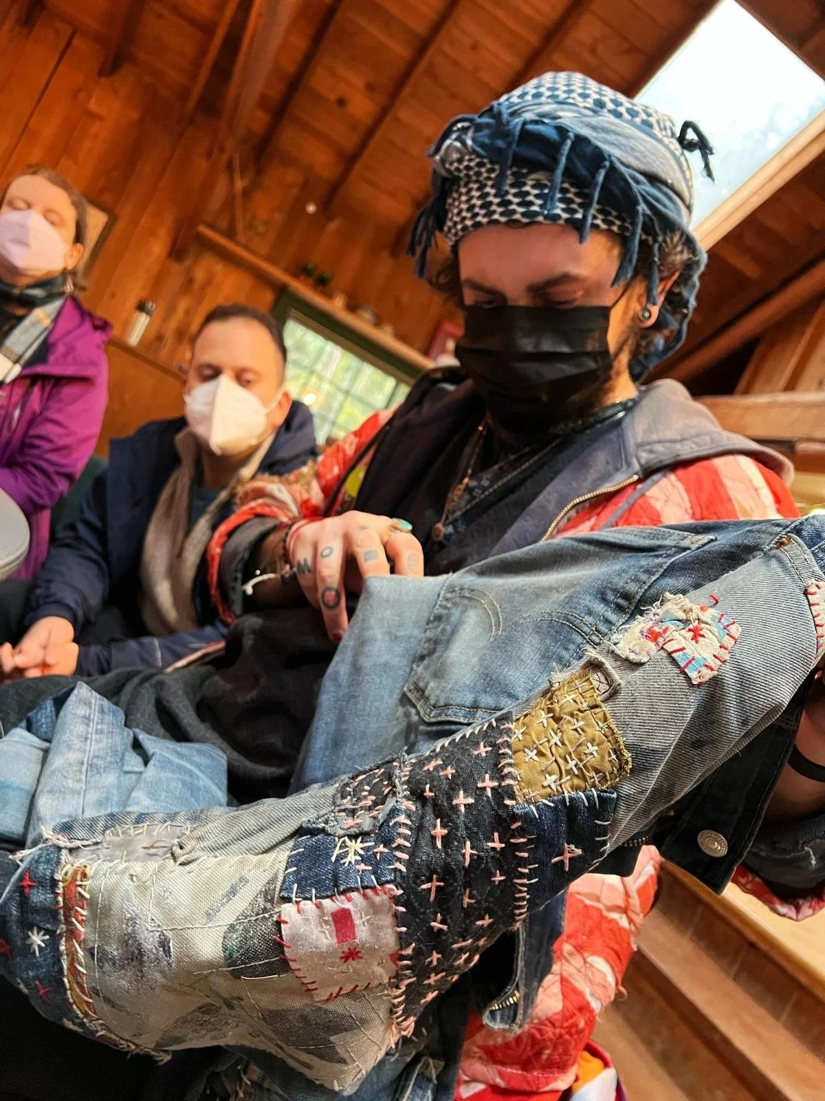 A person with tattoos on their arm is sitting at a table, showing their arm to others. They are wearing a black mask, a blue and white patterned headscarf, and a colorful, patchwork denim jacket. Two other people, also wearing masks, are seated nearb