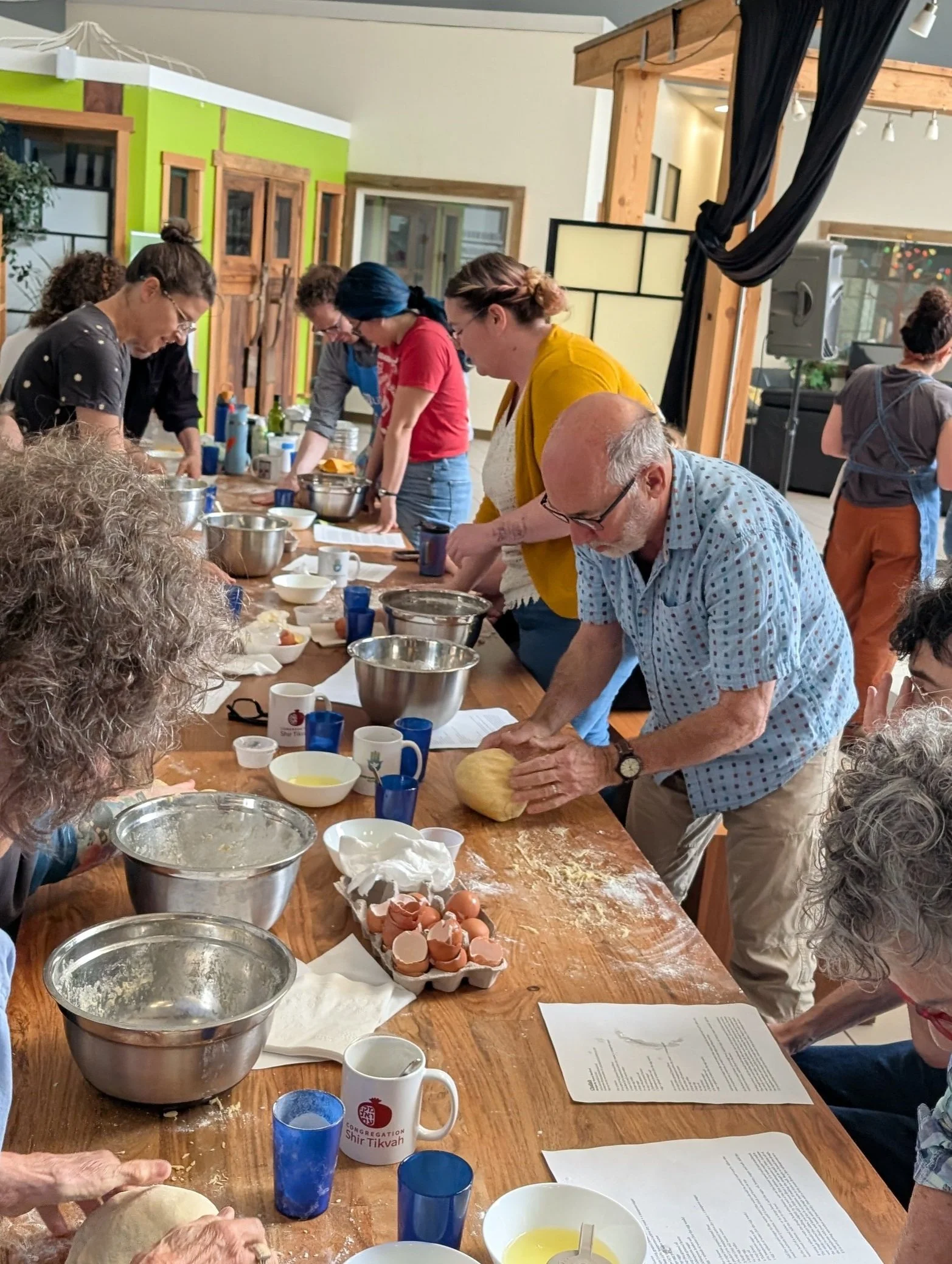 People participating in a baking class, kneading dough on a wooden table with baking ingredients and utensils, in a room with wooden beams and colorful walls.