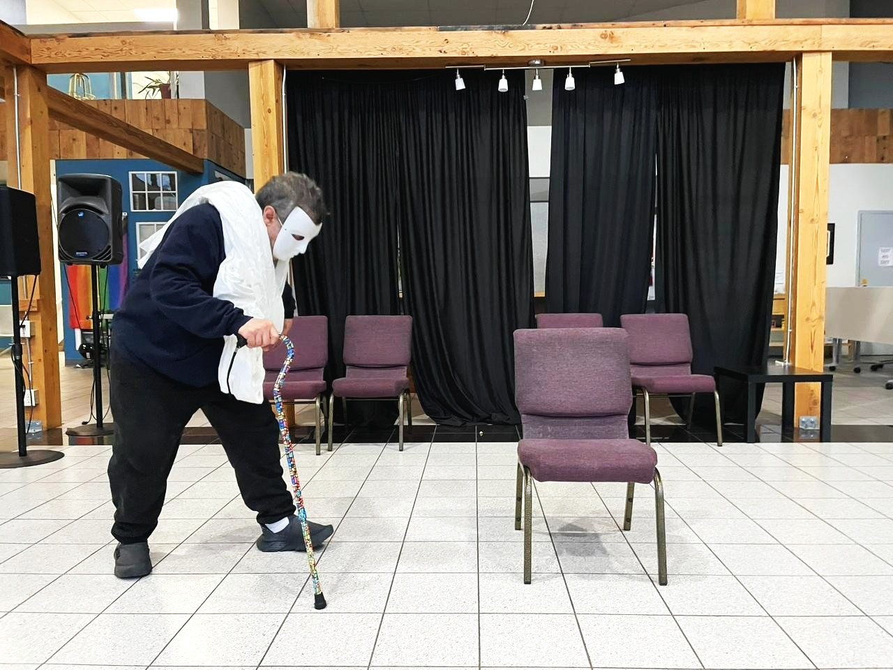 Person wearing a white mask and costume, sweeping a room with chairs on a tiled floor, stage with black curtains in the background.