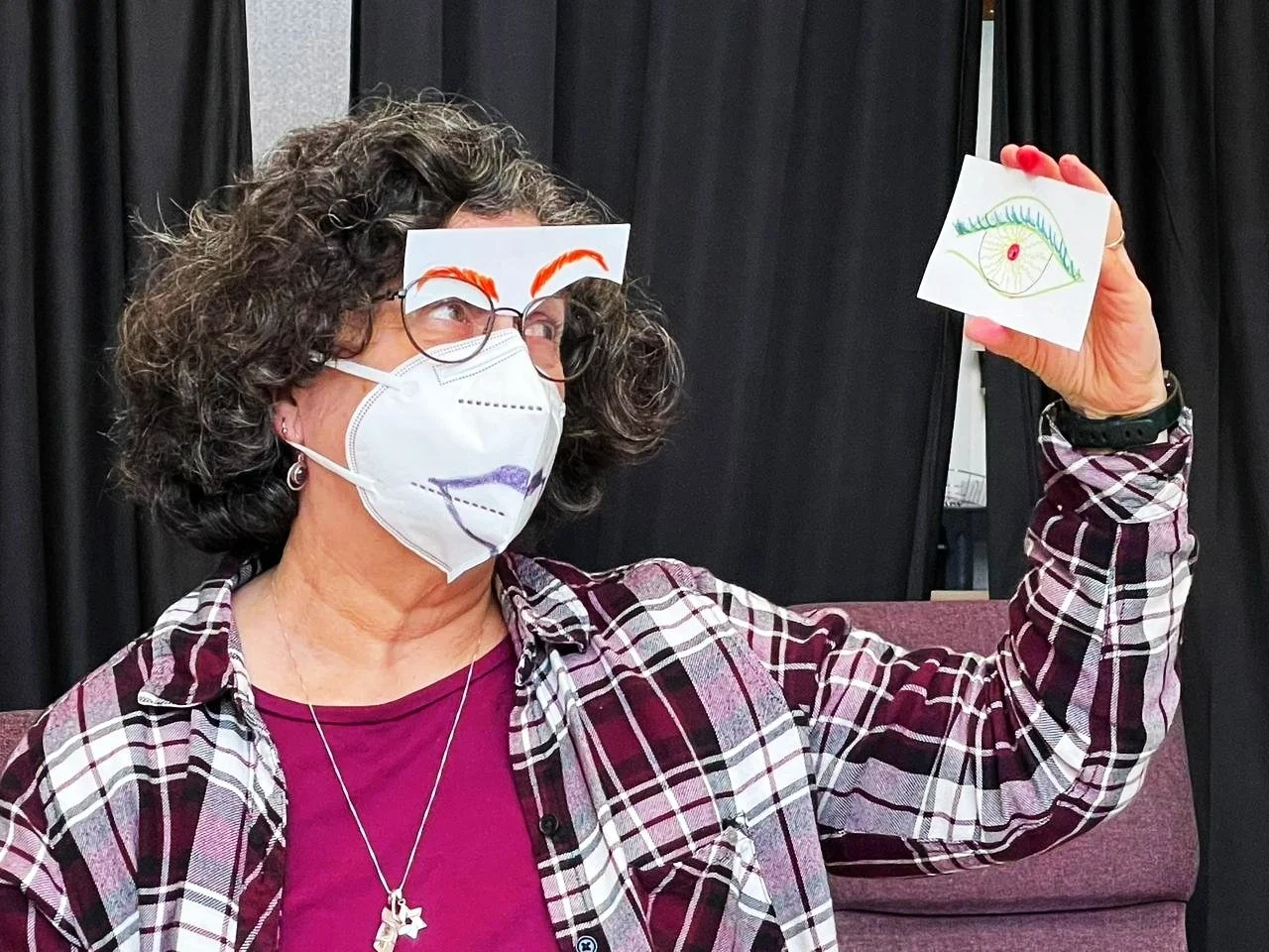 A woman wearing glasses and a face mask with colorful drawings, holding a paper with an eye drawing.