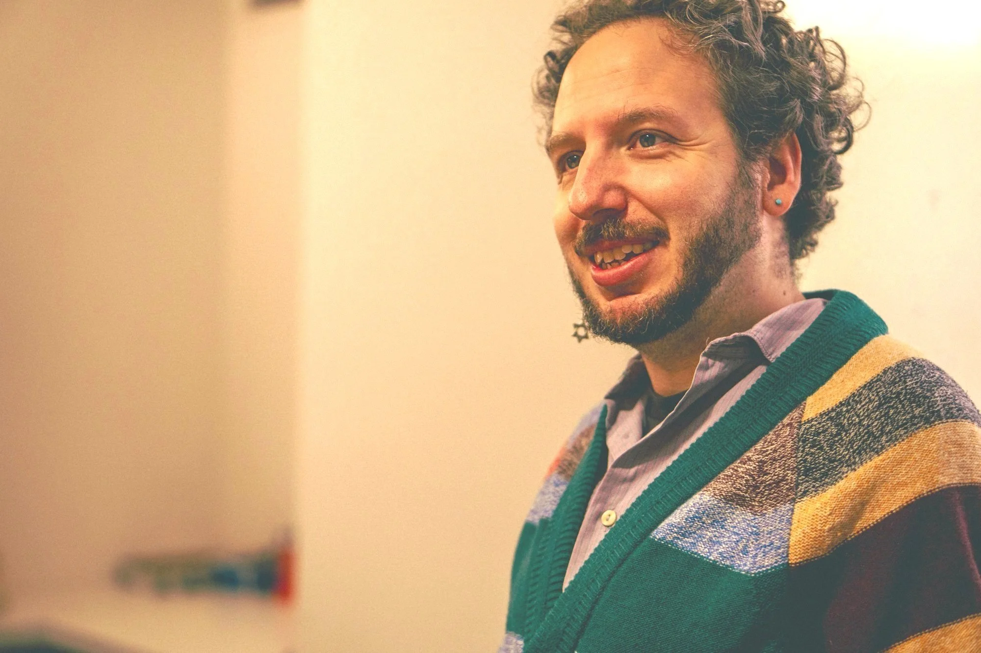 A man with curly hair, a beard, and earrings smiling in a room with a beige background, wearing a multicolored striped sweater over a light purple shirt.