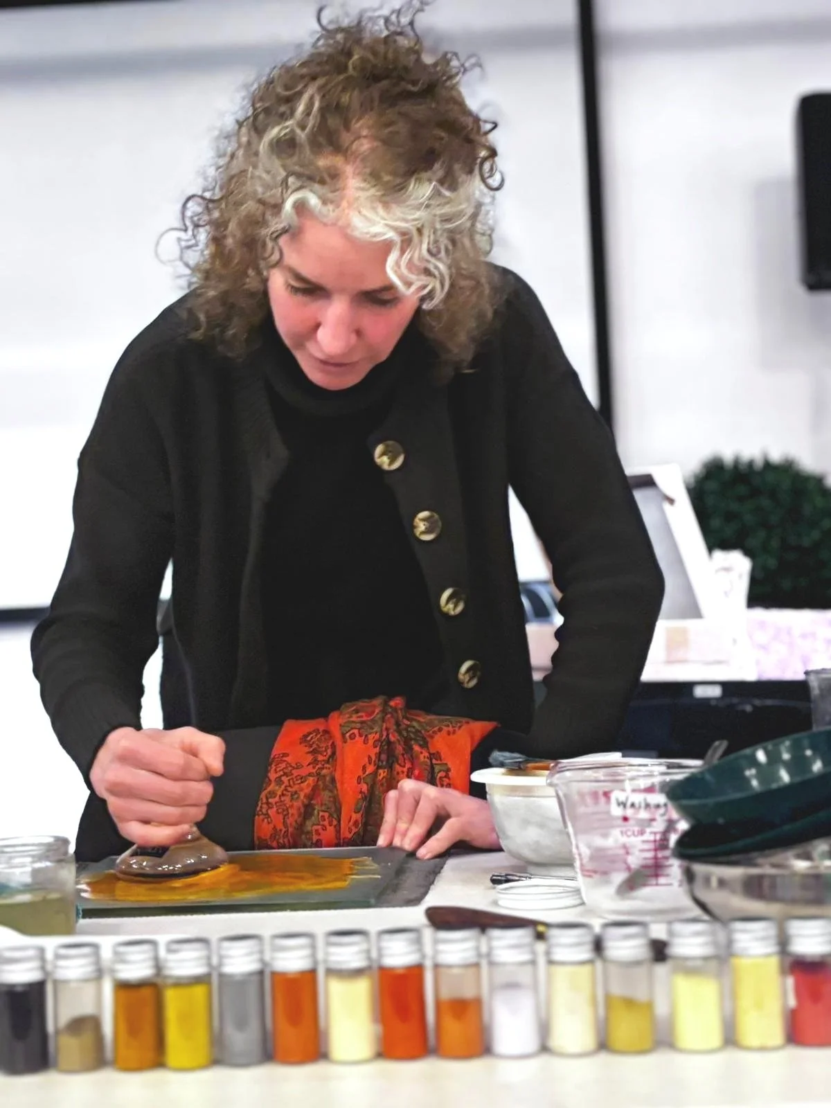 A woman with curly, gray hair, wearing a black jacket with gold buttons, is working on a craft project at a table.