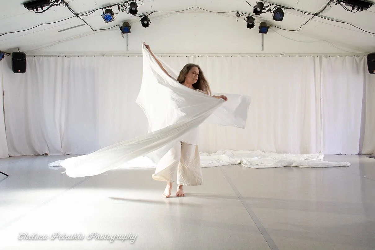 A woman dancing barefoot in a white flowing dress inside a white tent with curtains, stage lights, and speakers, holding a large white fabric.