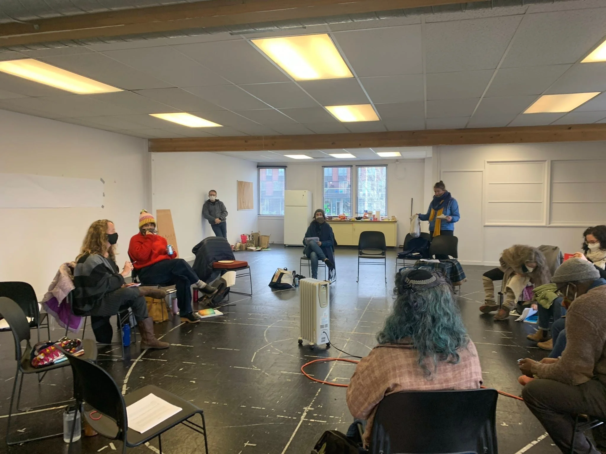 Group of people sitting in a semi-circle in a spacious room with large windows, some wearing masks, engaged in a meeting or workshop. There are chairs, a radiator, and some supplies on a table and the floor.