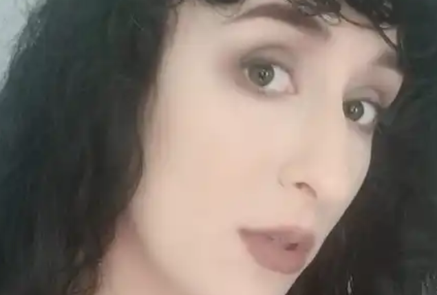 A person with fair skin, dark wavy hair, and makeup looking at the camera.