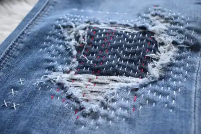 Close-up of a ripped and frayed hole in blue denim fabric with red stitching and faded edges.