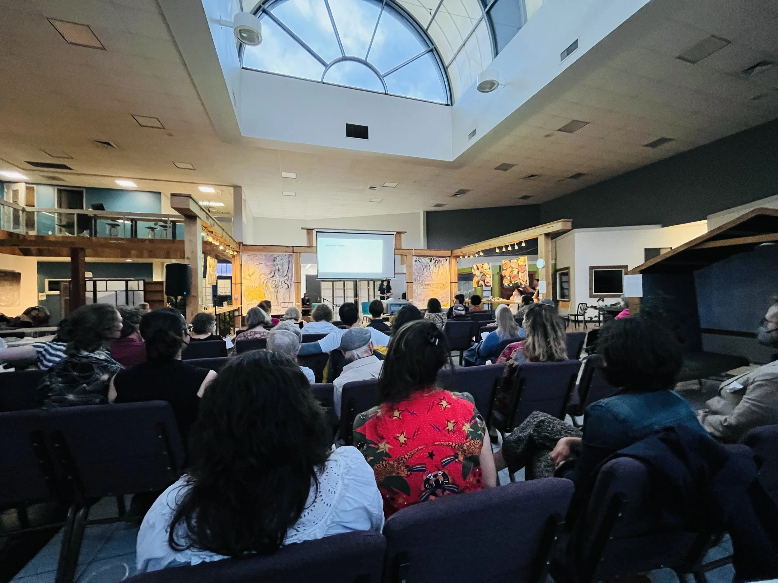 Audience seated facing a stage with a presenter and a projection screen inside a modern building with a glass skylight ceiling.