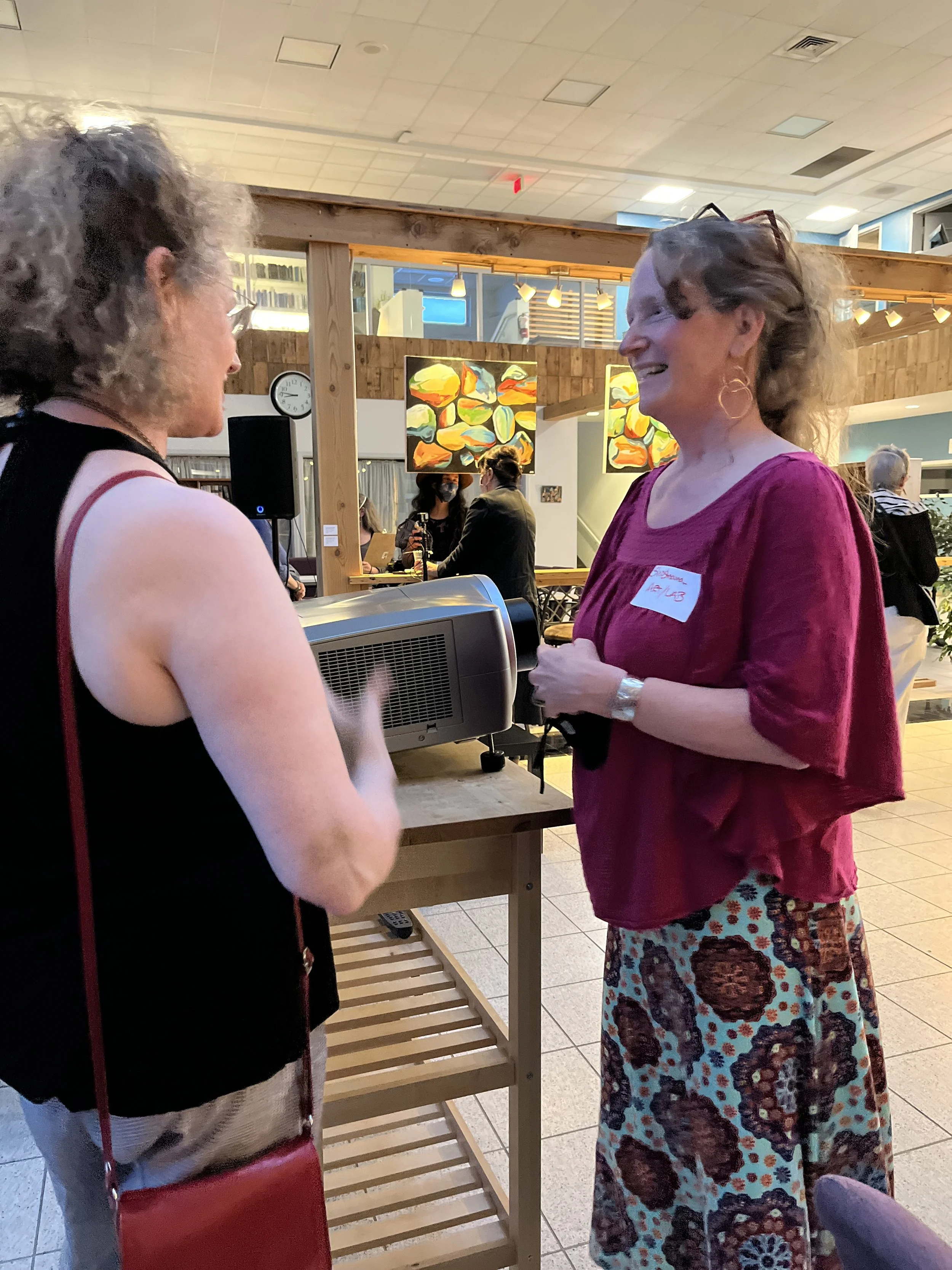 Two women are engaging in a conversation at a reception area, smiling and standing near a wooden table with a small electronic device on it. They are inside a brightly lit room with art on the walls.